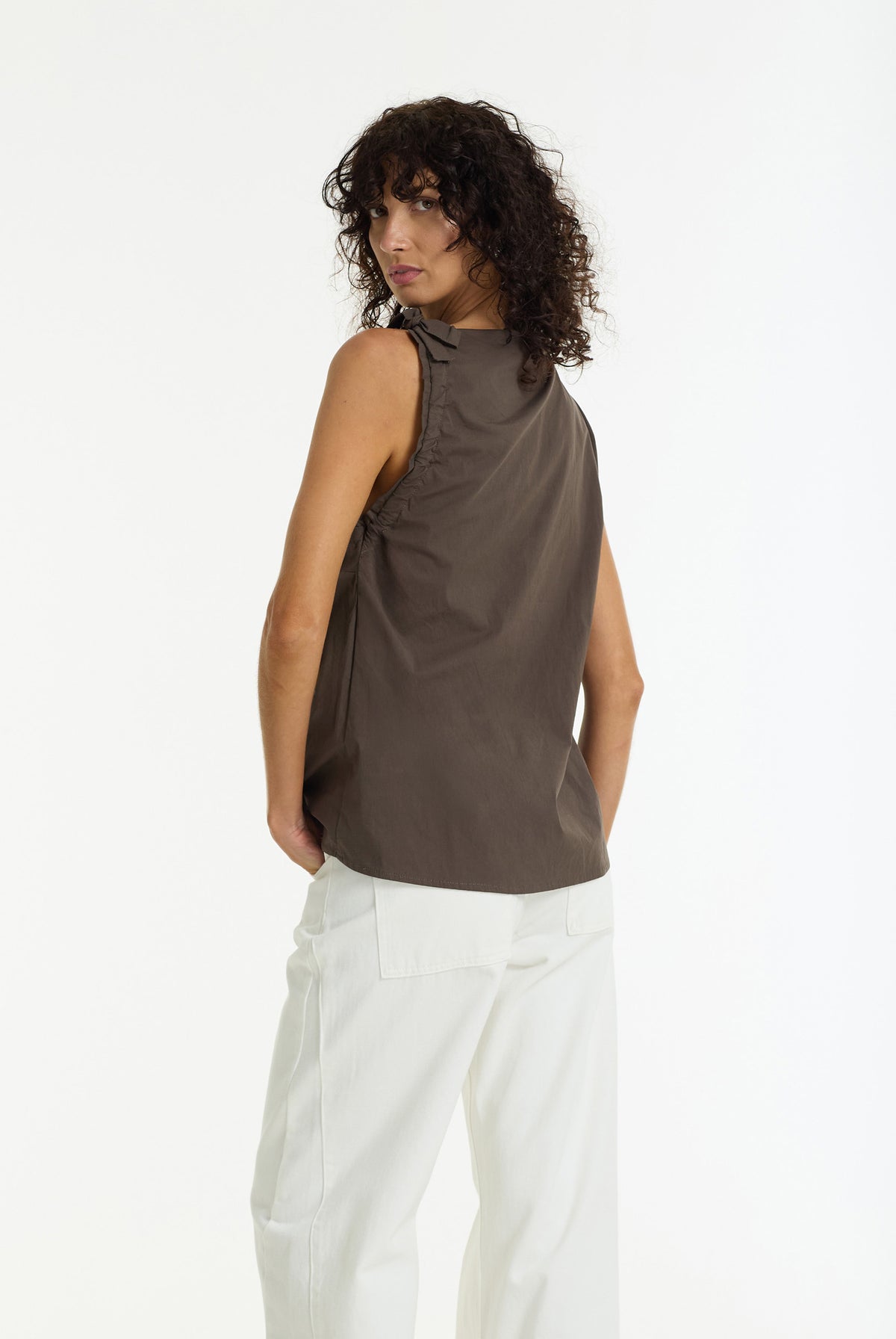 A model with curly dark hair is shown from a three-quarters back view, wearing a sleeveless, high-neck taupe-brown top and white trousers against a plain white studio background.