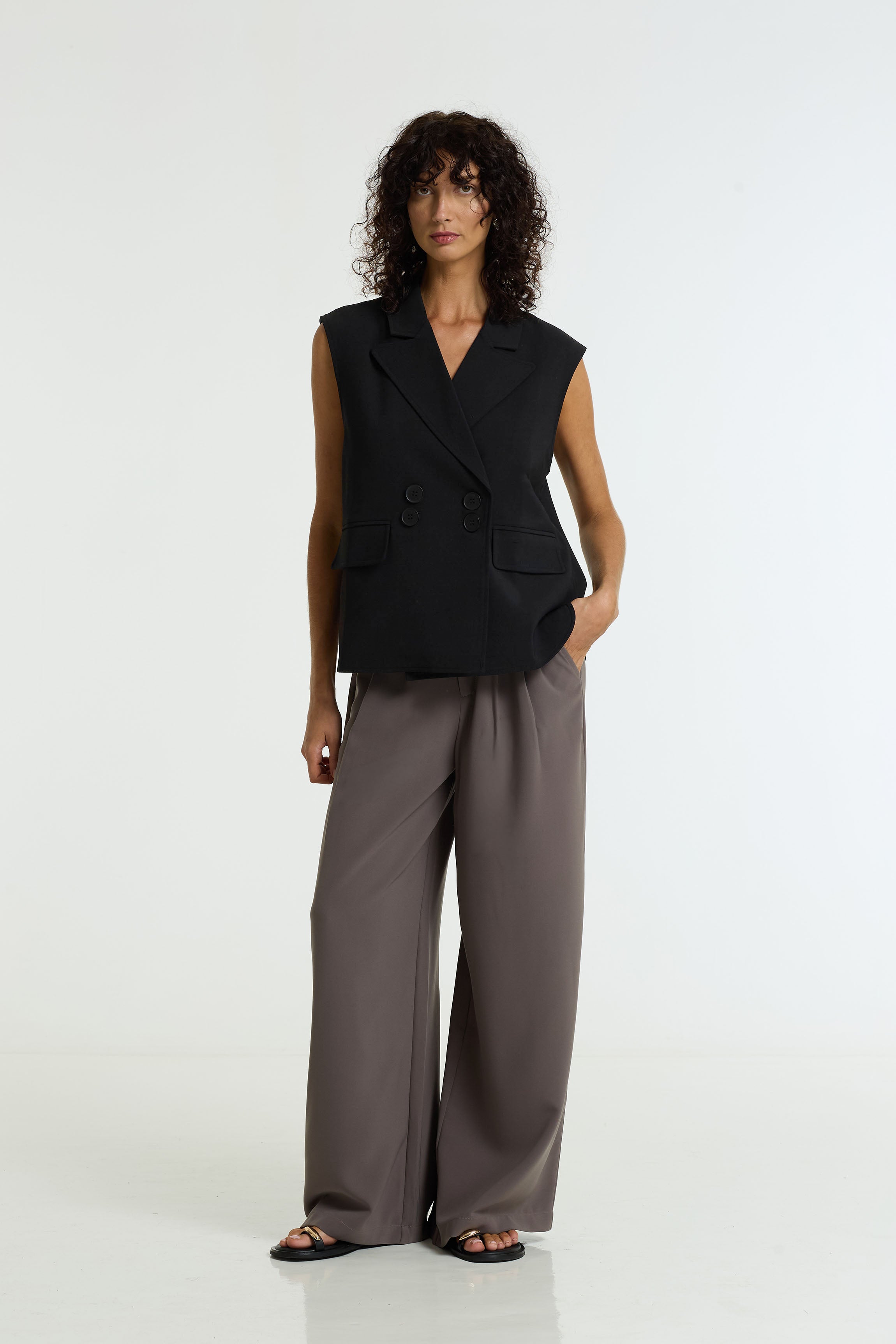 A model stands against a white studio background wearing a black sleeveless double-breasted blazer vest and the Marco Wide Leg Pant in charcoal / black by BLAK