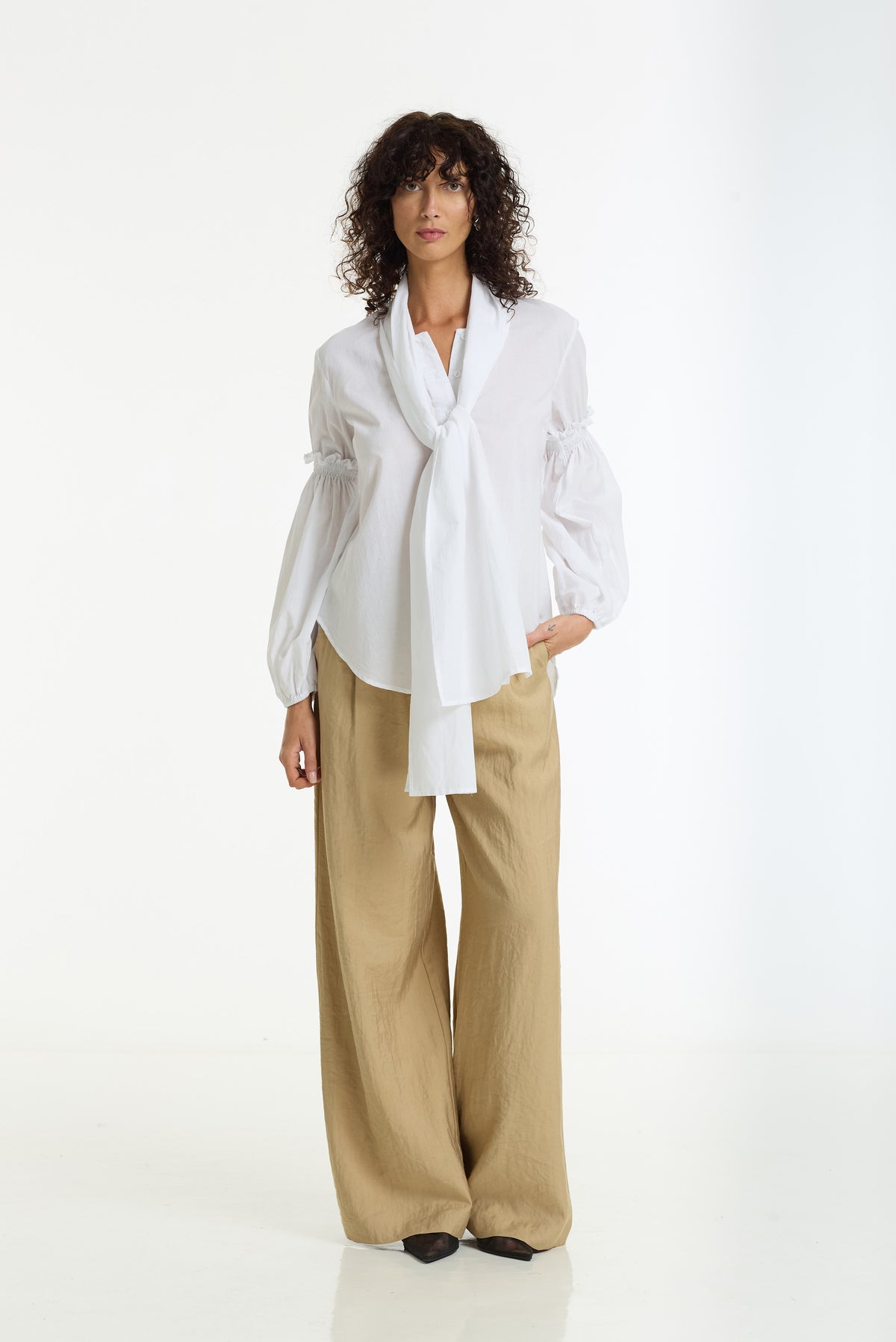 A model stands against a white studio background wearing a white long-sleeve blouse with a shawl collar and tie detail, paired with wide-leg beige trousers.