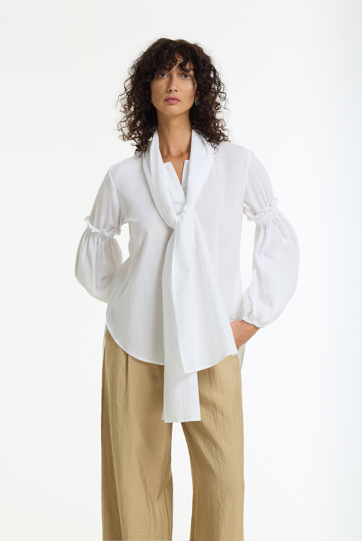 A woman modeling a white long-sleeve blouse with a draped shawl collar and gathered sleeves, paired with beige wide-leg trousers, against a plain white studio background.