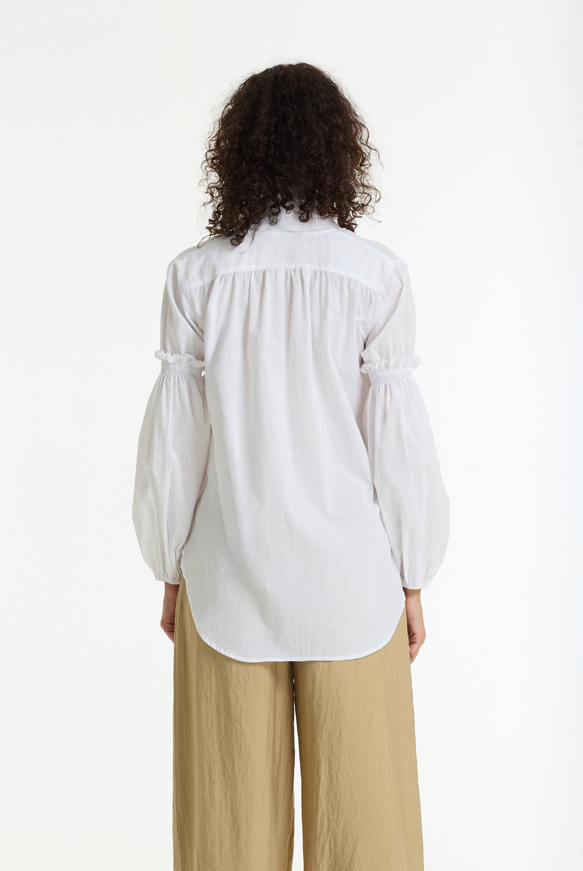 A woman viewed from the back wearing a white long-sleeve blouse with gathered puff sleeves and beige wide-leg pants, against a plain white studio background.