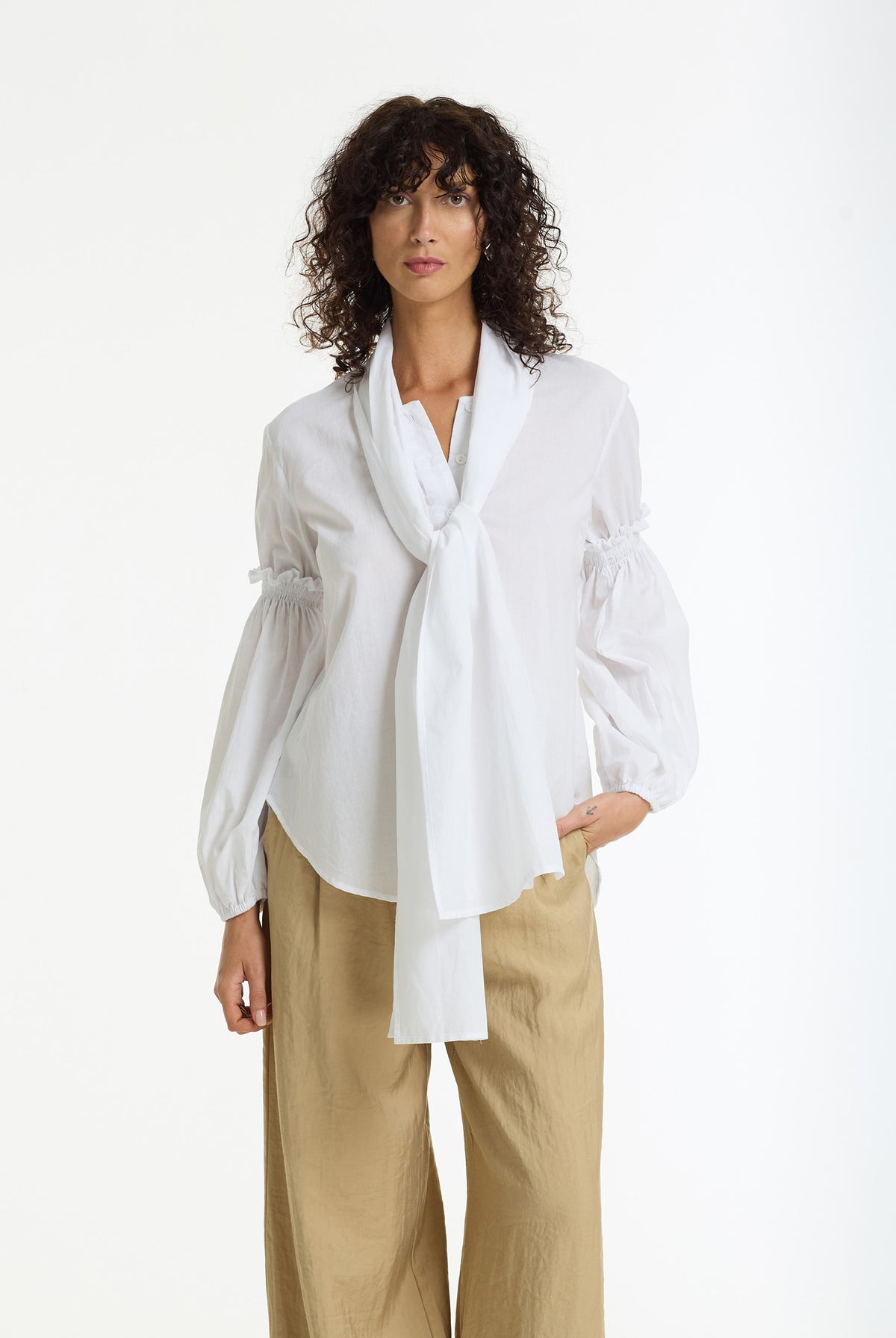 A model with curly dark hair wearing a Margo Tie Neck Cotton Voile Shirt in White by BLAK it's a white long-sleeve blouse with a draped tie-neck detail and gathered puff sleeves, paired with beige wide-leg trousers, against a plain white studio background.