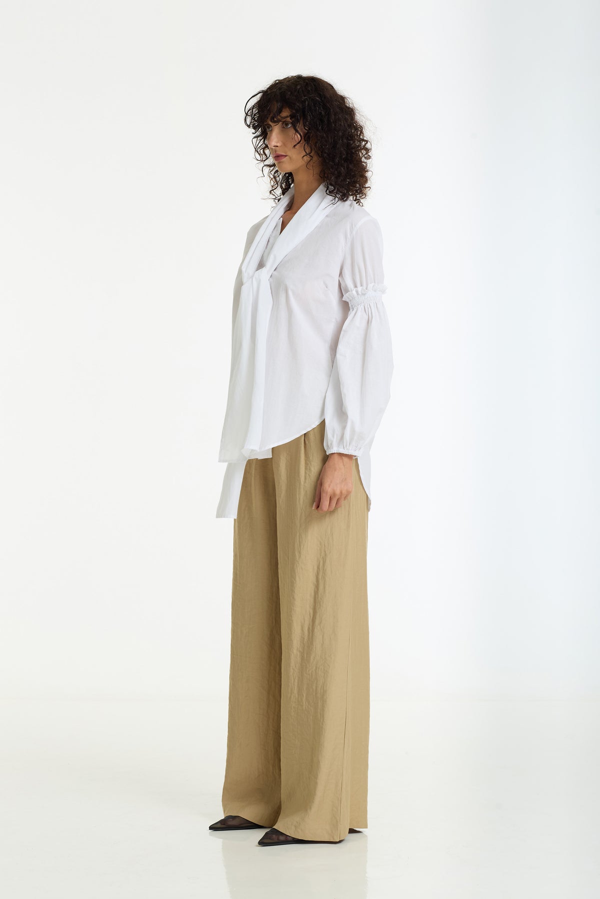 A woman stands in profile wearing a white long-sleeve blouse with gathered sleeves and a tie neckline, paired with wide-leg tan trousers, against a clean white studio background.