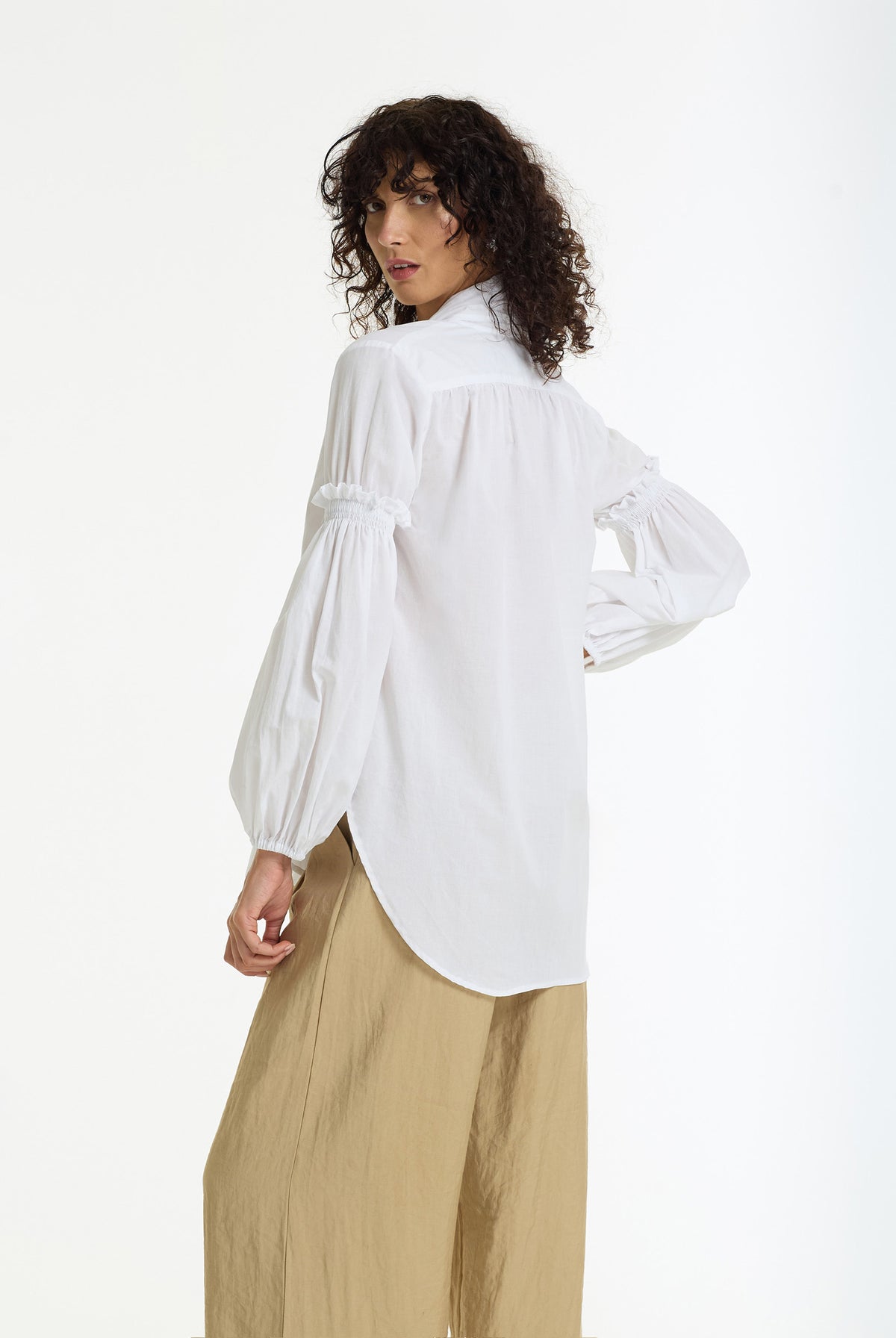 A model wearing a white long-sleeve blouse with gathered puff sleeves and a curved hem, paired with beige wide-leg trousers, photographed against a plain white studio background.