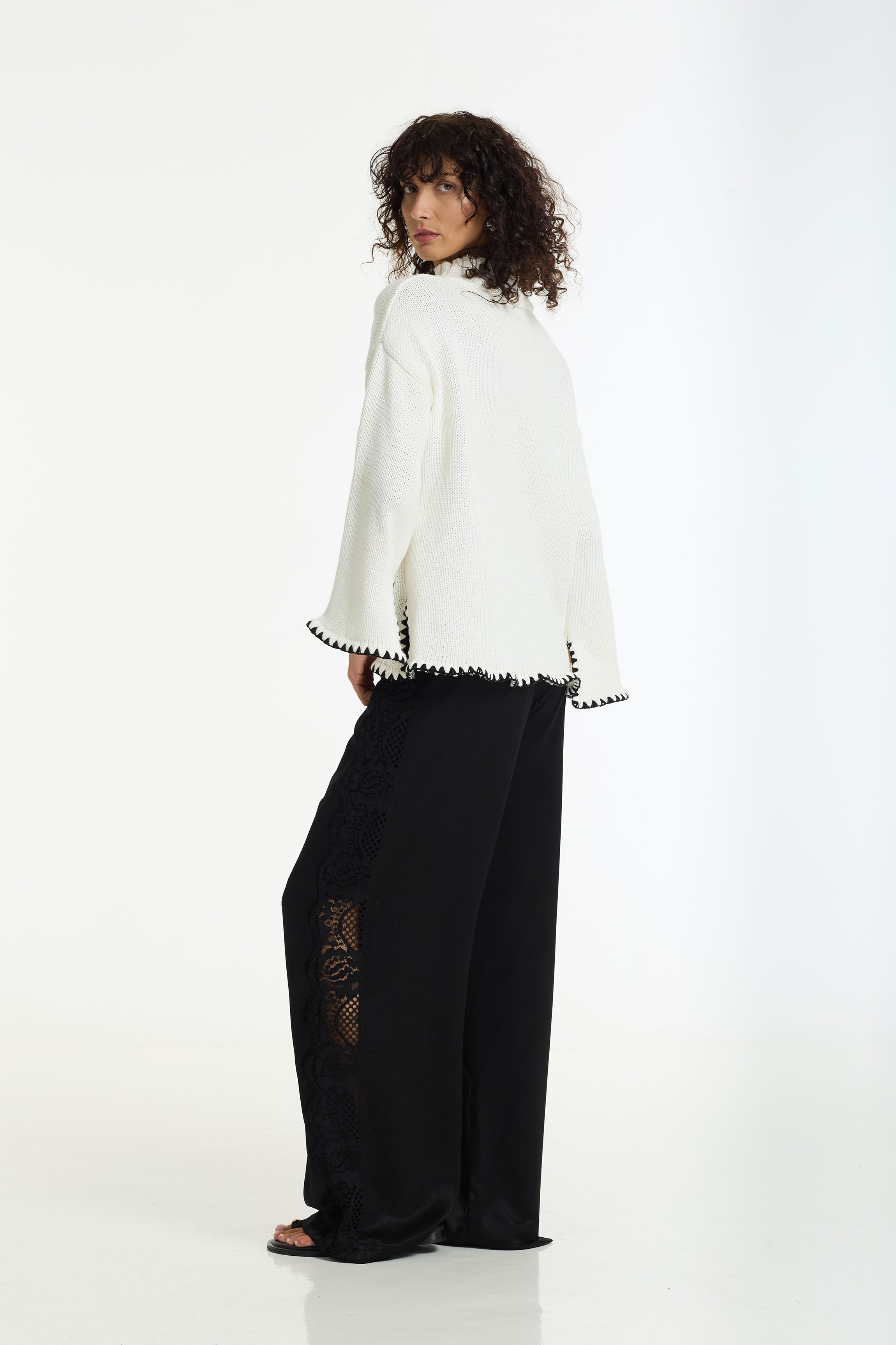 A model is shown from the back wearing a White Vogue Sweater with black blanket stitch edging and the Marlow Satin Lace Pant in black by BLAK, set against a plain white studio background.