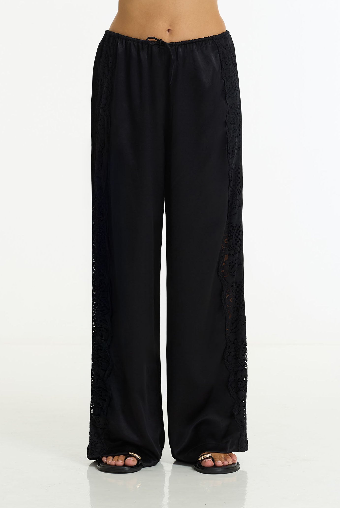 Marlow Satin Lace Pant in Black by BLAK, shown on a model from the waist down wearing black sandals against a white studio background.