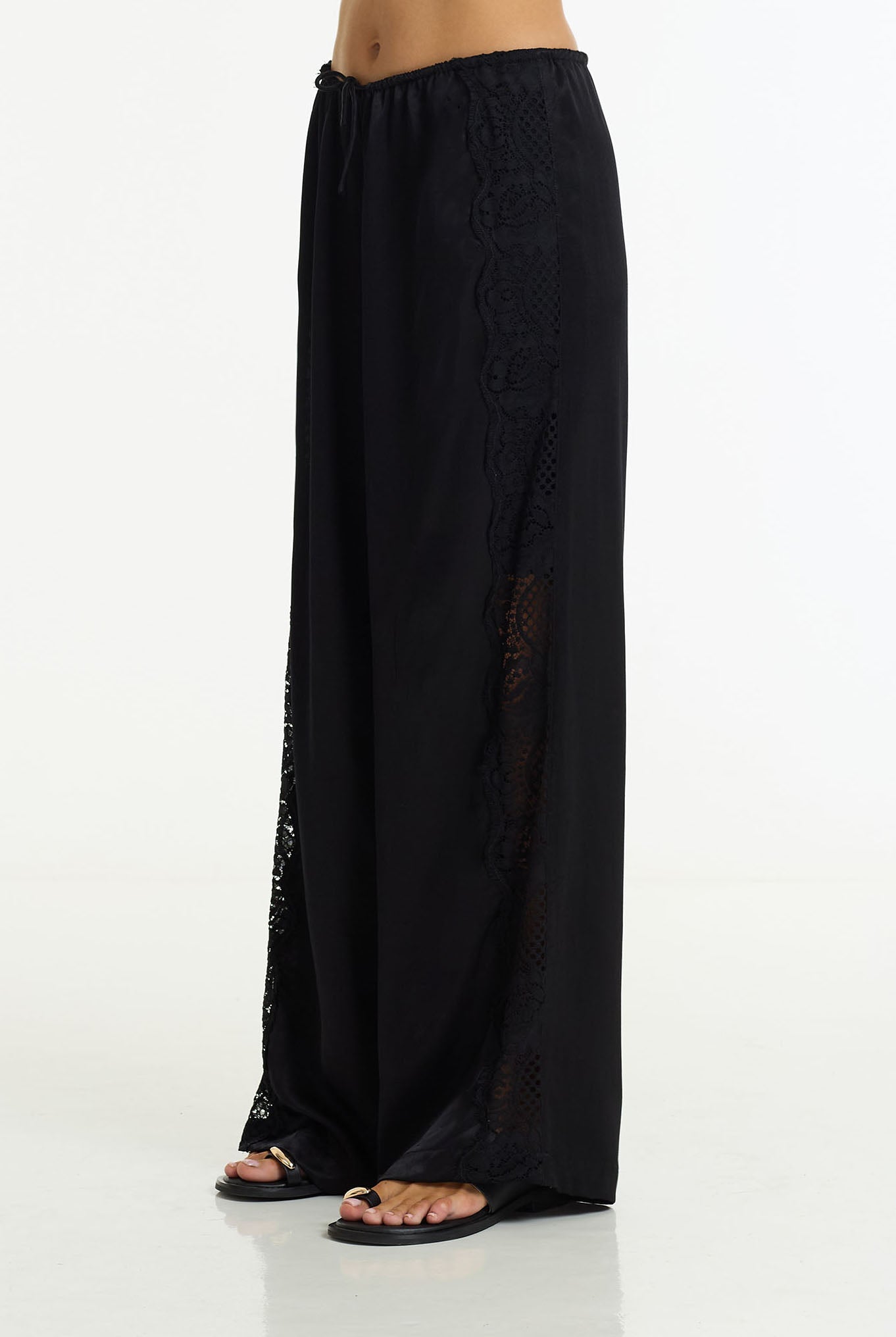 Black wide-leg pants with lace side panels and a drawstring waist, shown on a model from the waist down against a white studio background.