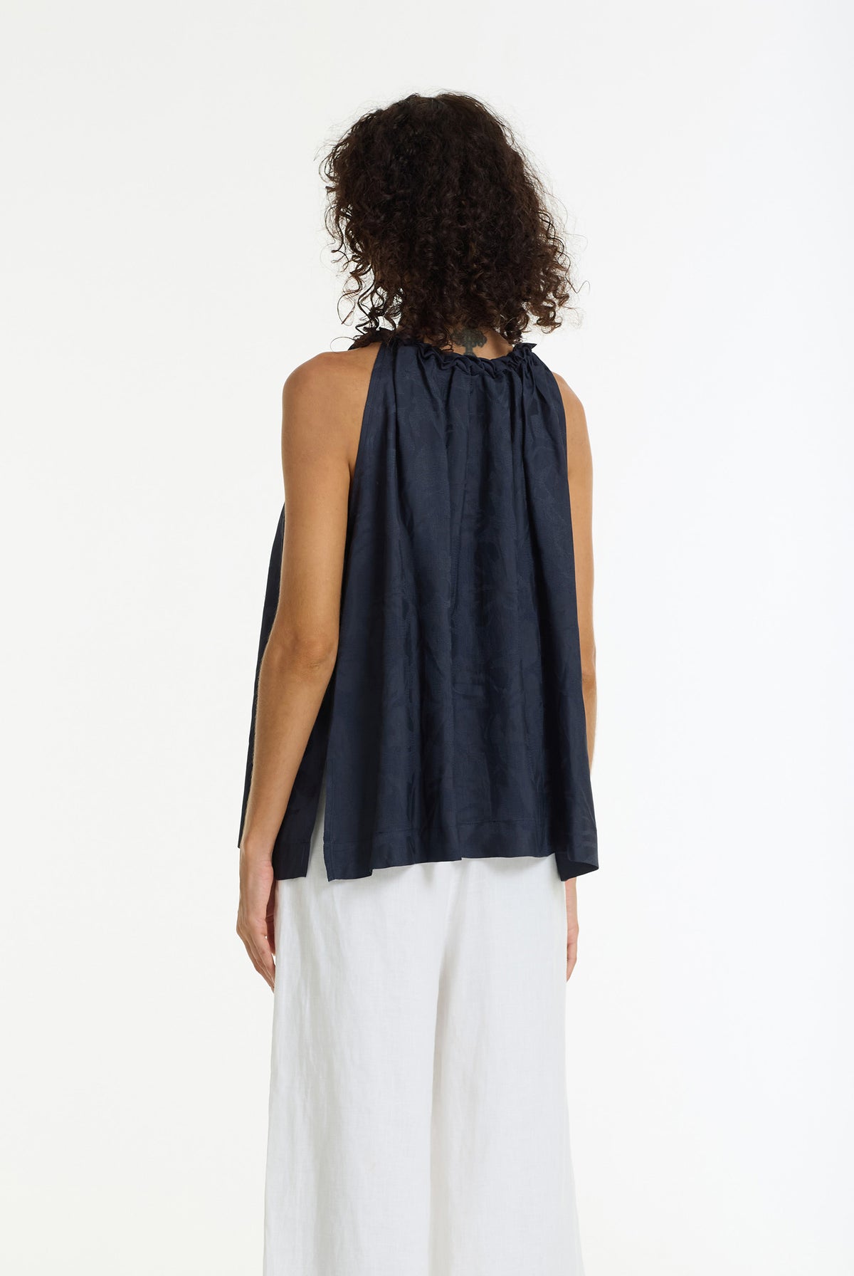 A model shown from the back wearing a sleeveless navy blue top with a gathered neck and white wide-leg pants, set against a plain white studio background.