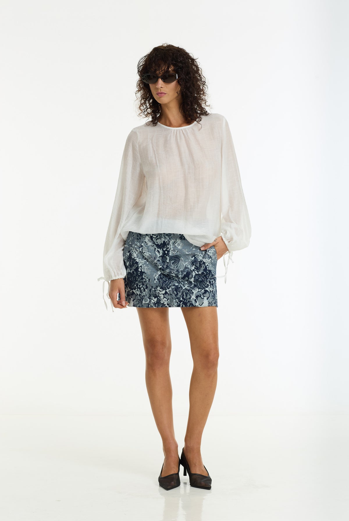 A model wearing a sheer white long-sleeve blouse with tie details at the cuffs and a Nocturne Floral Mini Skirt in Dark Denim and Ivory by BLAK, paired with black high heels, against a plain white studio background.