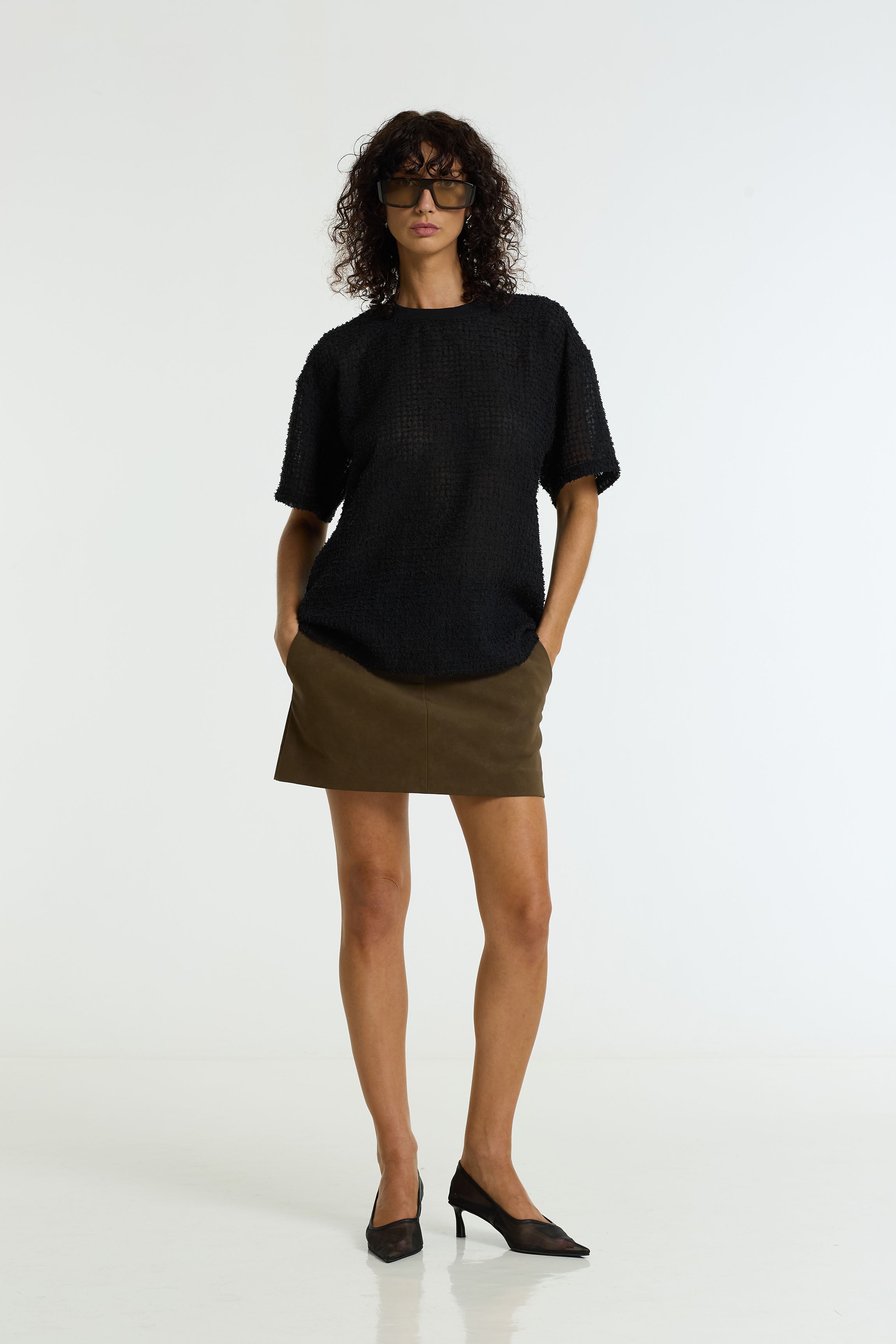 A model wearing a black short-sleeve knit top and a Nocturne Vegan Suede Mini Skirt in dark tan by BLAK, paired with black pointed-toe heels, photographed against a seamless white studio background.
