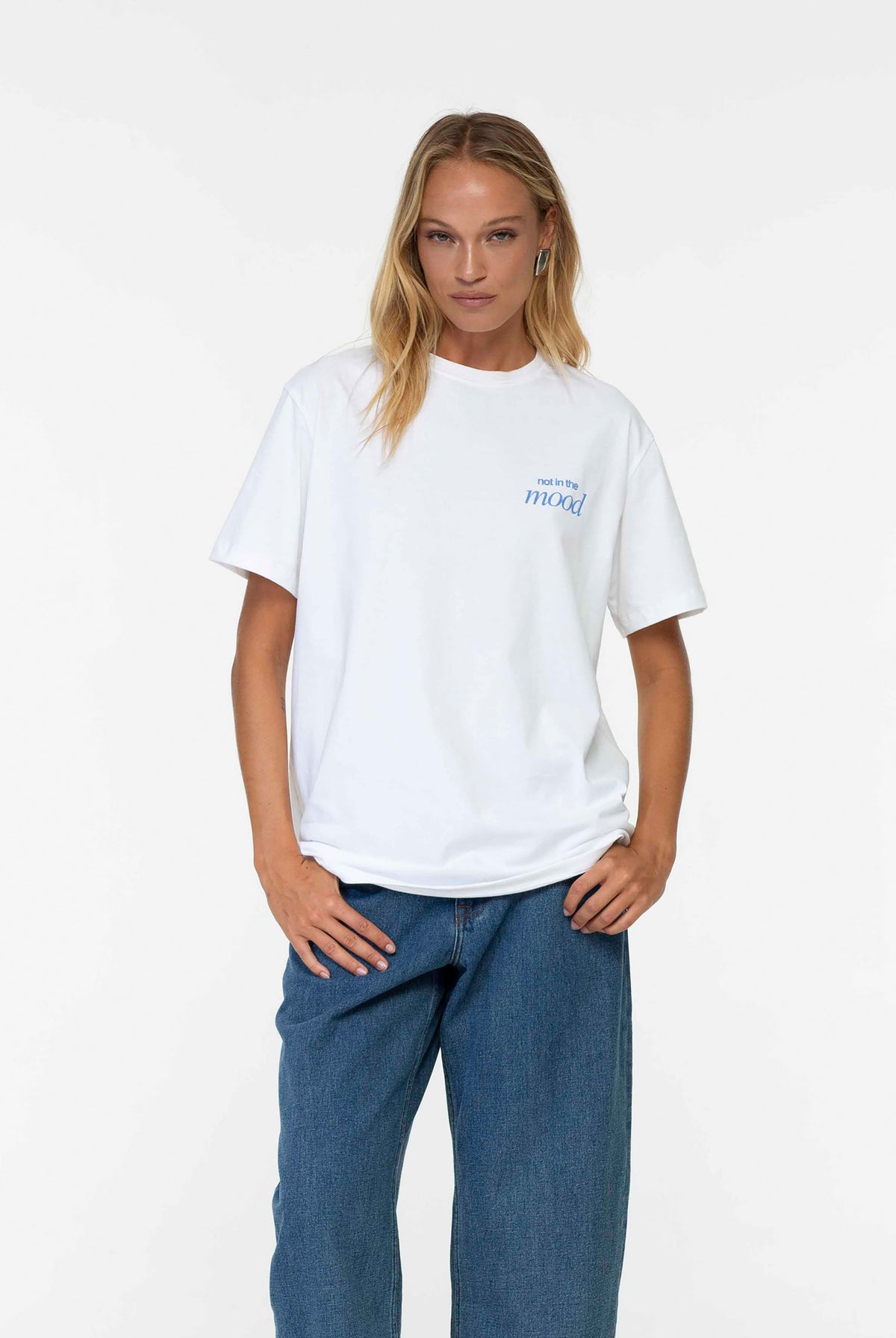 A woman modeling a white short-sleeve t-shirt with a small blue graphic on the left chest, paired with blue jeans, against a plain white studio background.