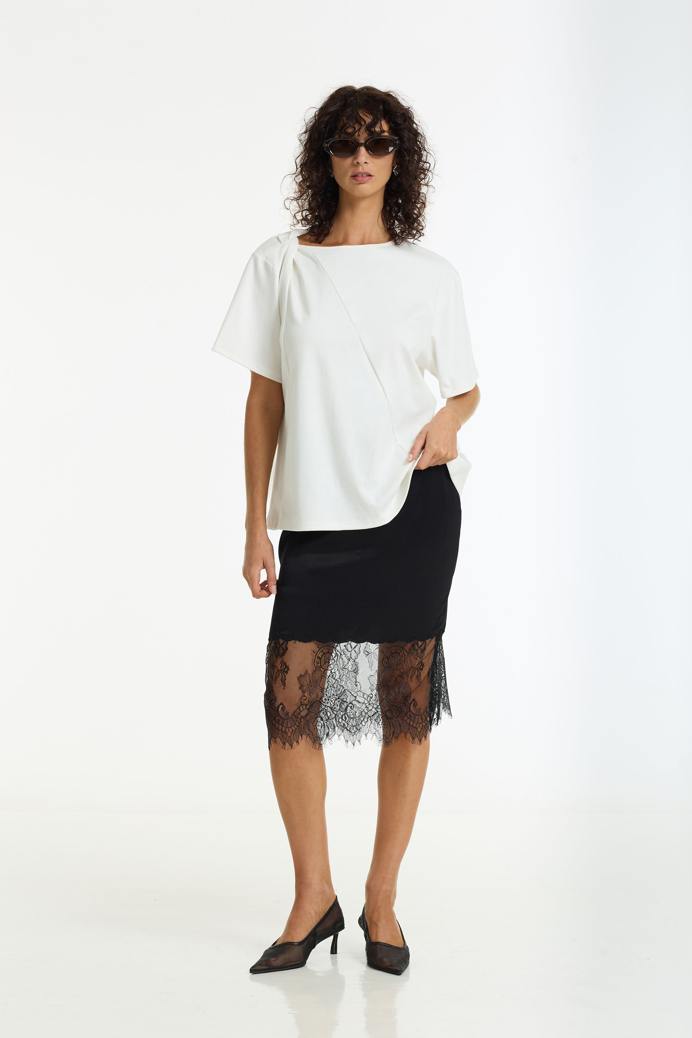 A model wearing a white short-sleeve top with a boat neckline and a Parisian Girl Satin Lace Skirt in black by BLAK styled with black pointed-toe heels against a white studio background.