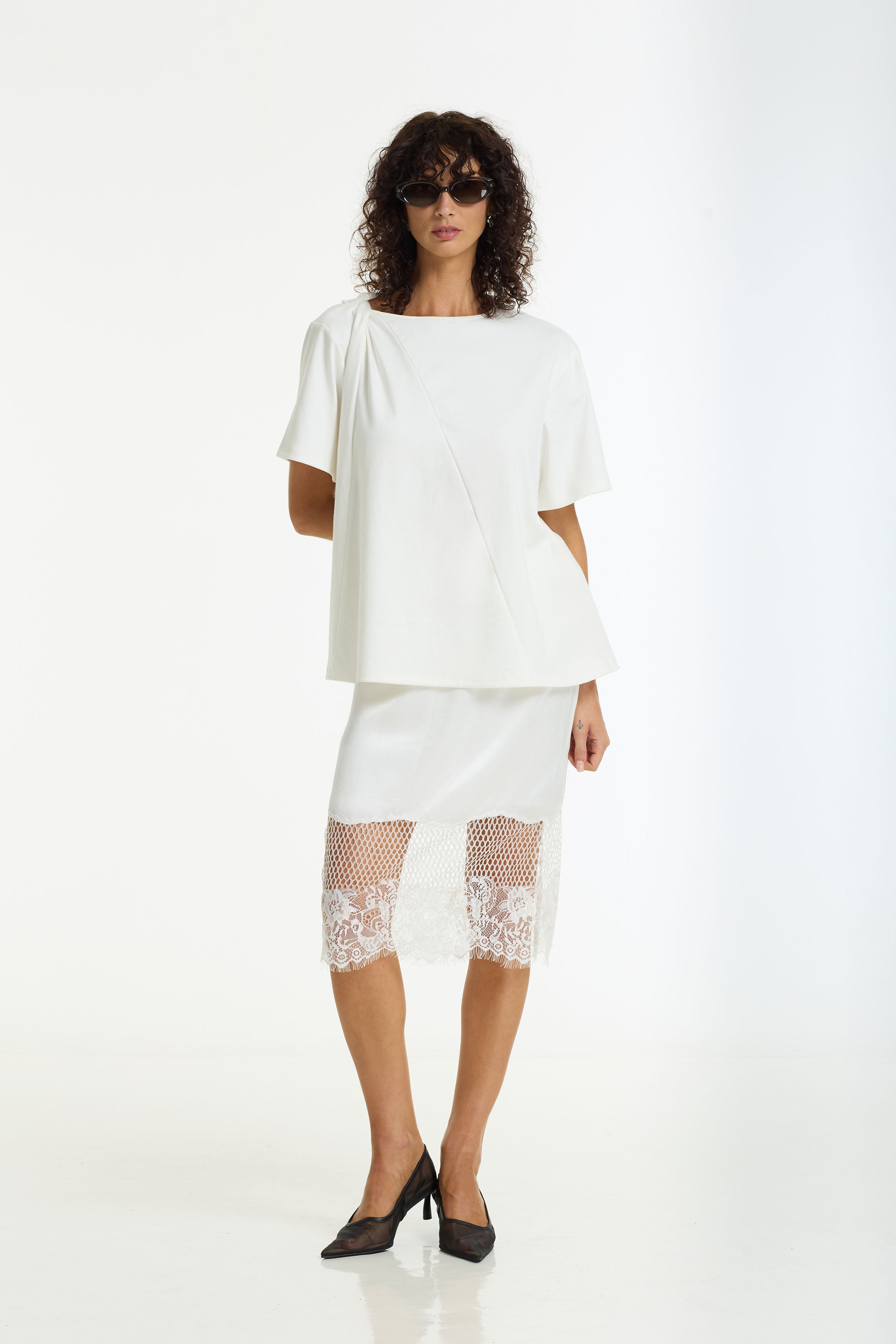 A model wearing a white short-sleeve top with draped side details and a Parisian Girl Satin Lace Skirt in ivory by BLAK, styled with black pointed-toe heels against a plain white studio background.