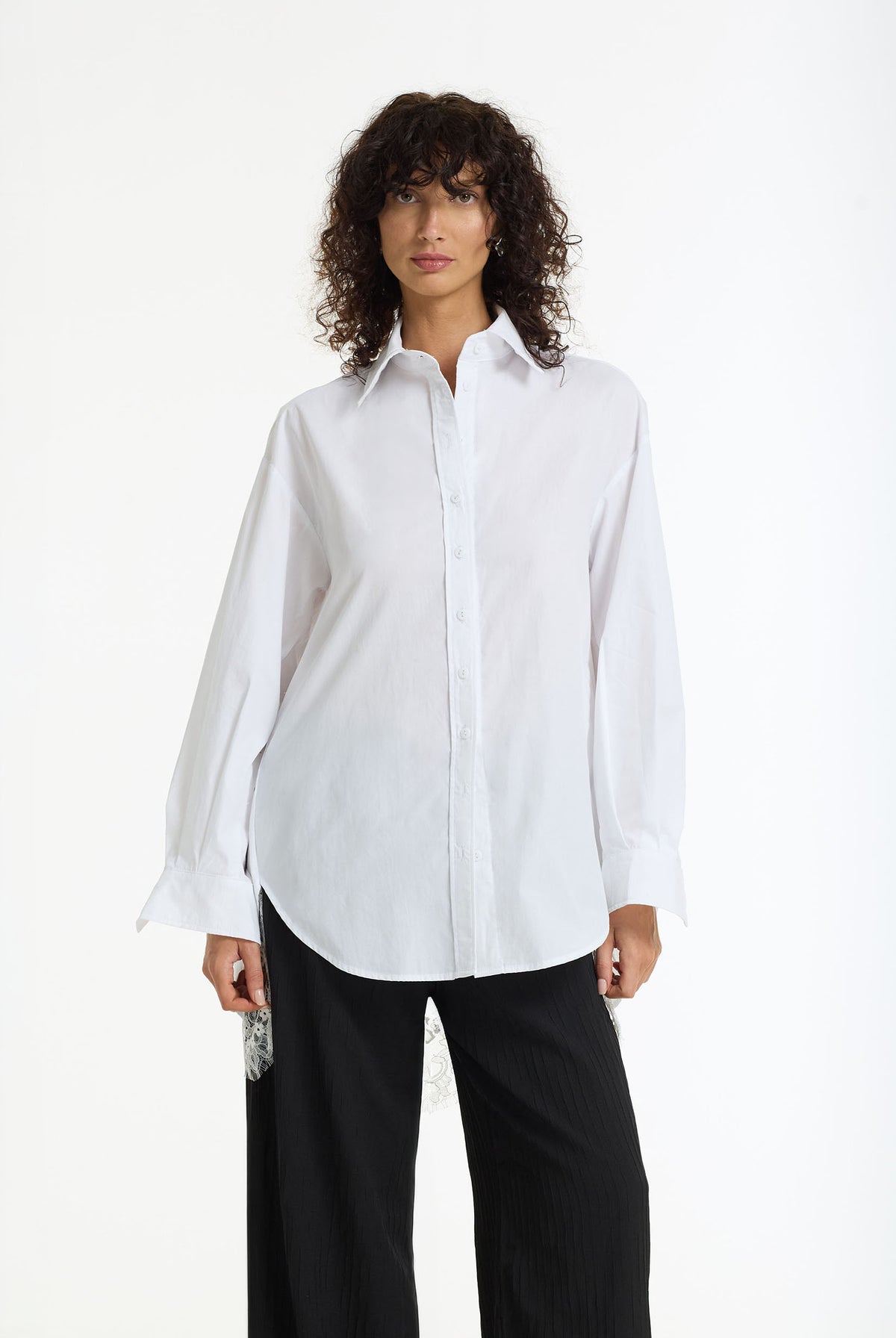 A woman modeling Reveal Lace Back Cotton Shirt, a white long-sleeve button-up shirt with a relaxed, oversized fit and black wide-leg trousers, shot against a plain white studio background.