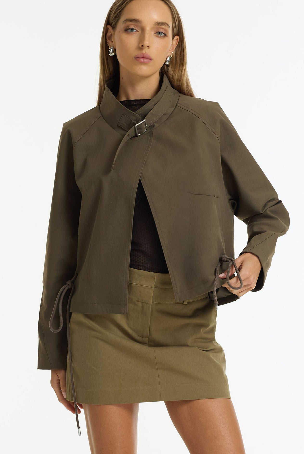 A model wearing an olive green cropped jacket with a unique asymmetrical collar and tie detail, paired with a matching olive green mini skirt and black top, against a plain white studio background.