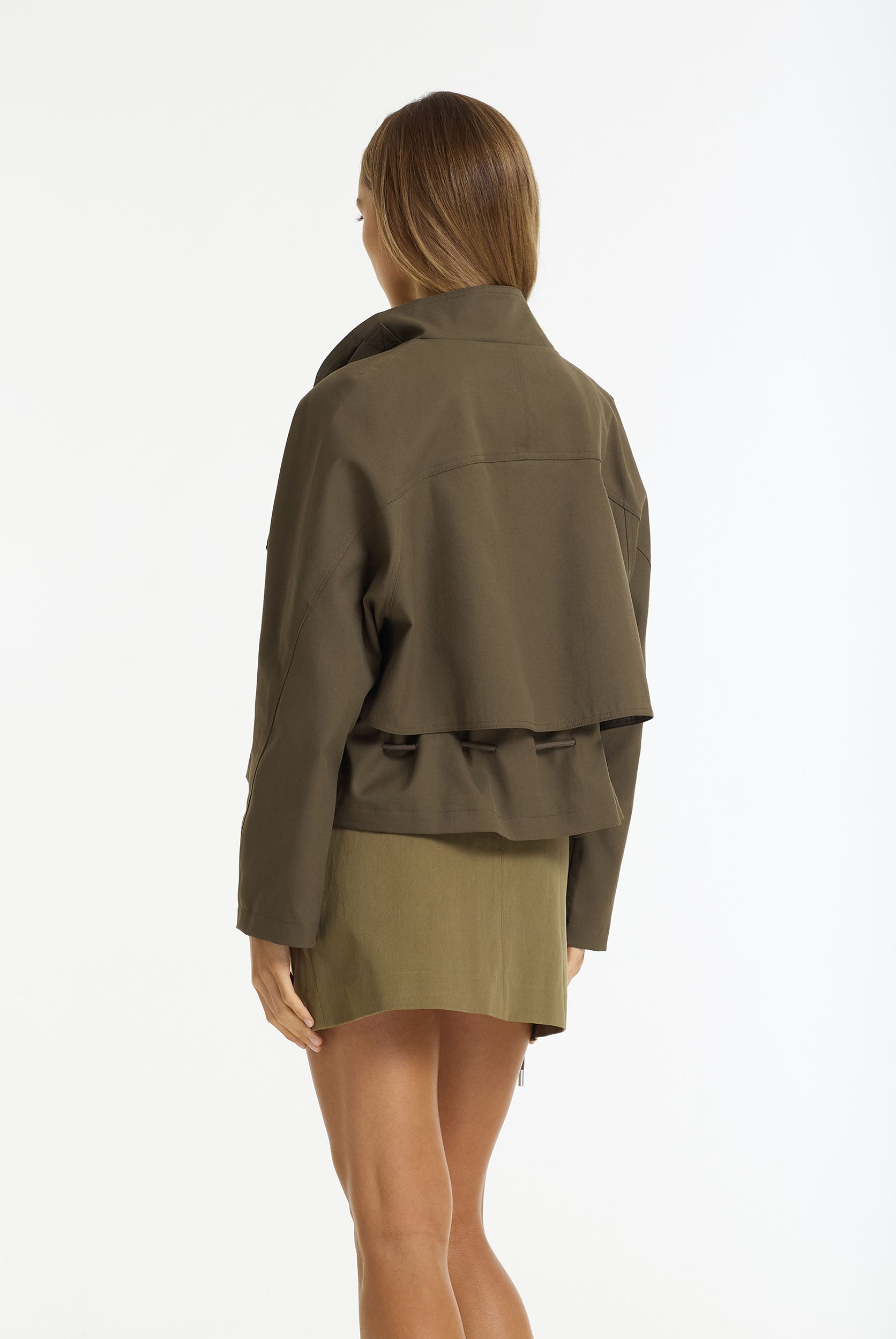 A woman shown from the back wearing a Rhodes Cropped Utility Jacket in dark khaki by BLAK and a olive green mini skirt, against a plain white studio background.