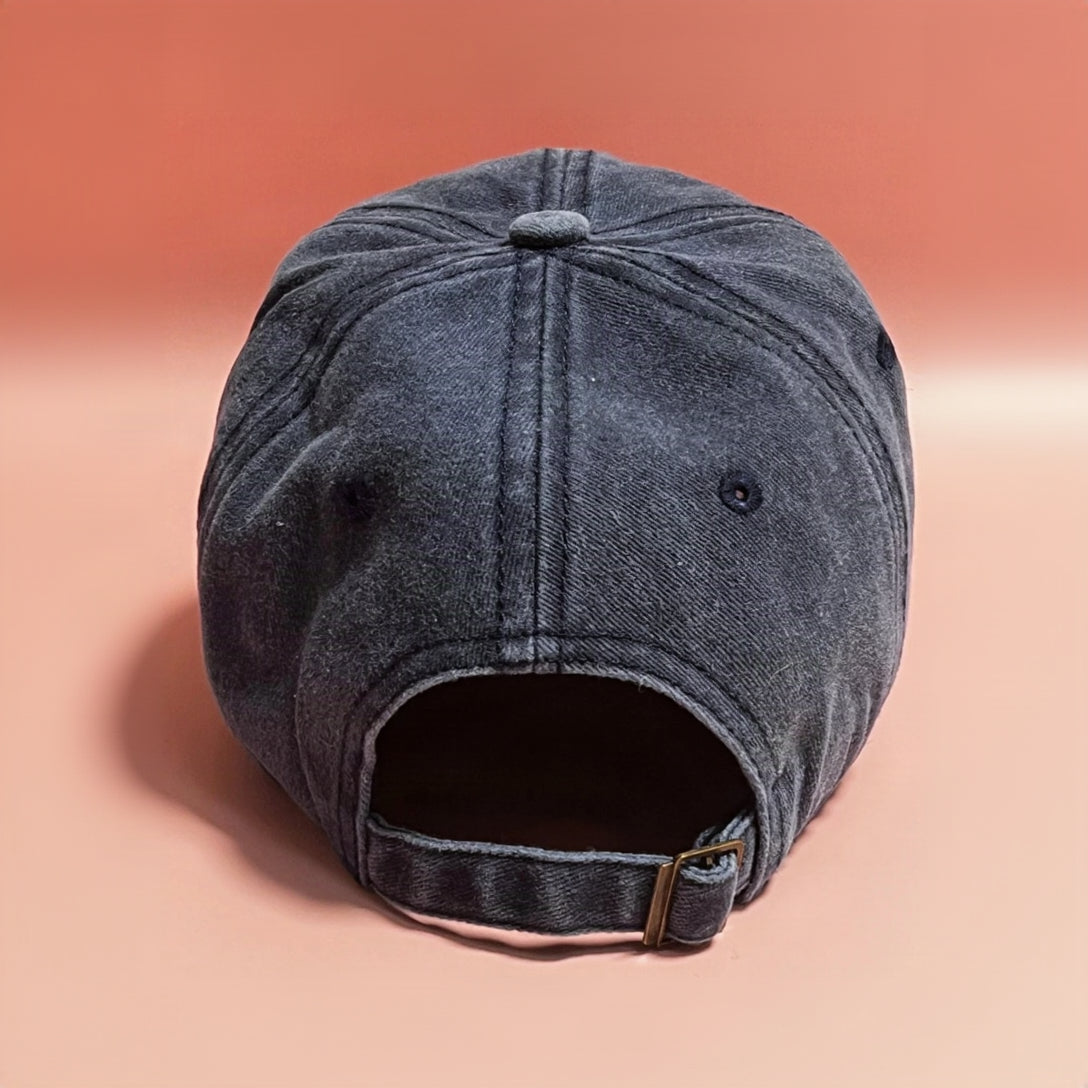 A dark gray washed cotton baseball cap shown from the back, featuring a curved back strap with a small gold-tone metal buckle, set against a solid peach-pink background.