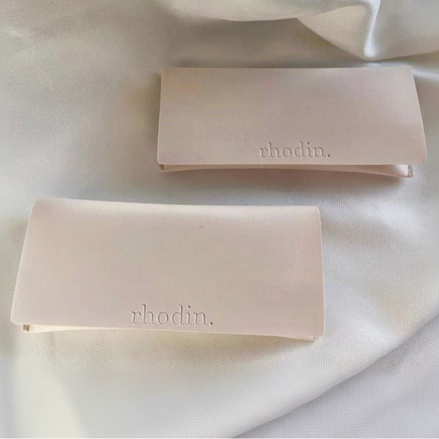 Two pale blush pink rectangular Rhodin eyeglass cases resting on draped white fabric, with the rhodin. logo debossed on each case.