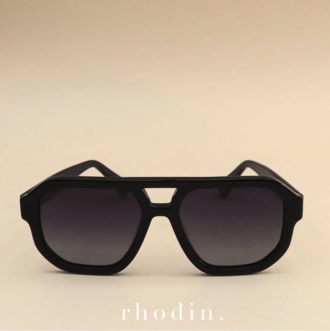 Black oversized geometric sunglasses with dark lenses, photographed against a warm beige gradient background. The brand name 'rhodin.