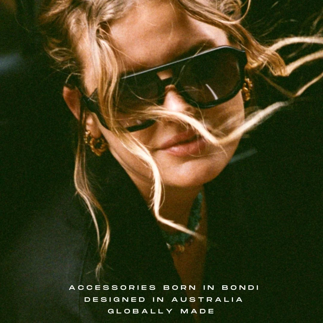 A close-up lifestyle shot of a woman with blonde hair blowing in the wind, wearing black sunglasses and gold hoop earrings. Text at the bottom reads 'ACCESSORIES BORN IN BONDI DESIGNED IN AUSTRALIA GLOBALLY MADE'.