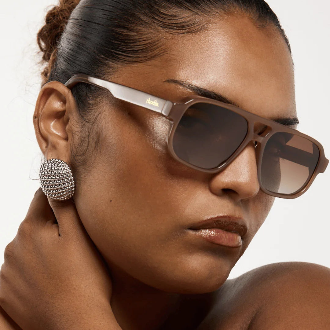 Woman wearing sunglasses with 'rhodin.' branding on a neutral background