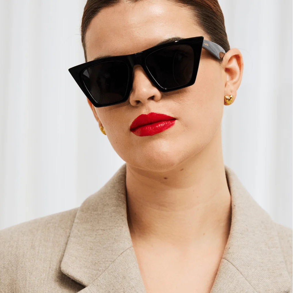 A close-up of a model wearing chunky black cat-eye sunglasses, a beige blazer, and red lipstick against a soft white curtain background. The brand name 'rhodin' is printed at the bottom.