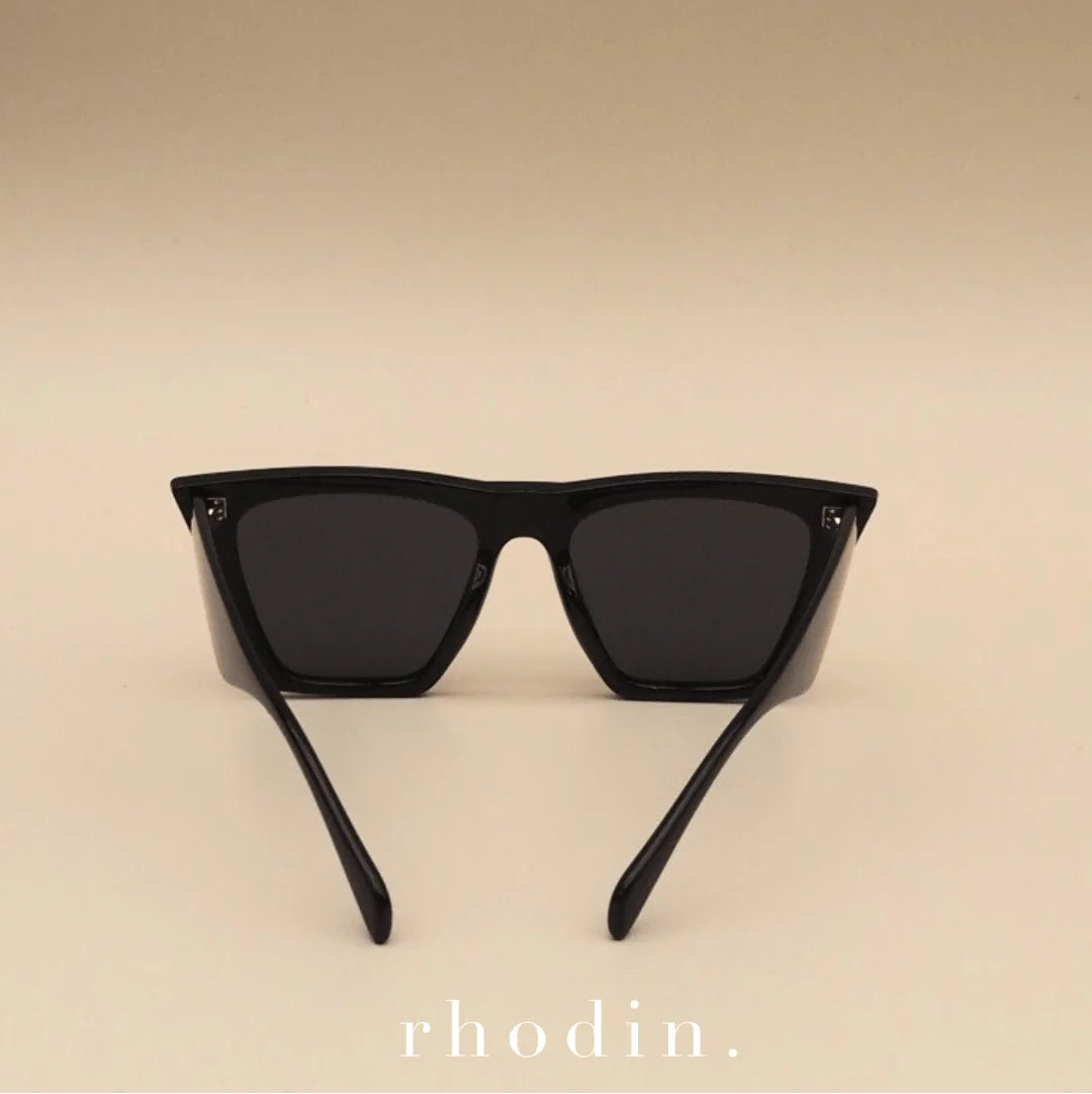 Black square sunglasses with dark lenses and thin temples, photographed against a warm beige gradient background. The word 'rhodin.