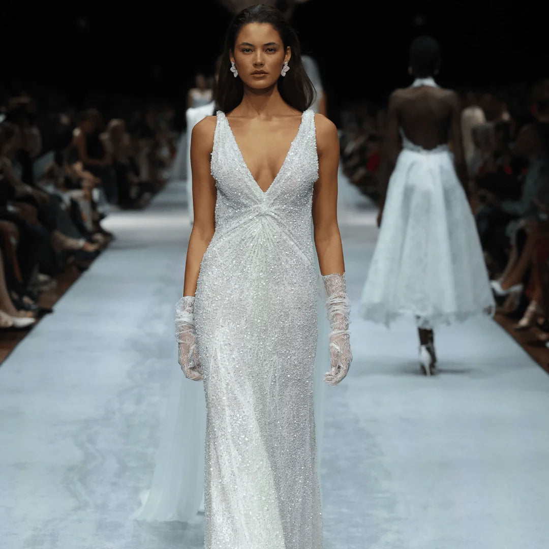 A model walks down a fashion runway wearing a floor-length white gown with a deep V-neckline, fitted silhouette, and intricate beaded texture. She is also wearing long white gloves.
