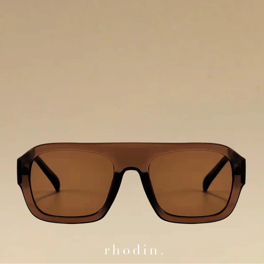 Rhodin oversized square sunglasses with a translucent dark brown acetate frame and matching brown lenses, shown against a solid beige background with the rhodin. logo at the bottom.
