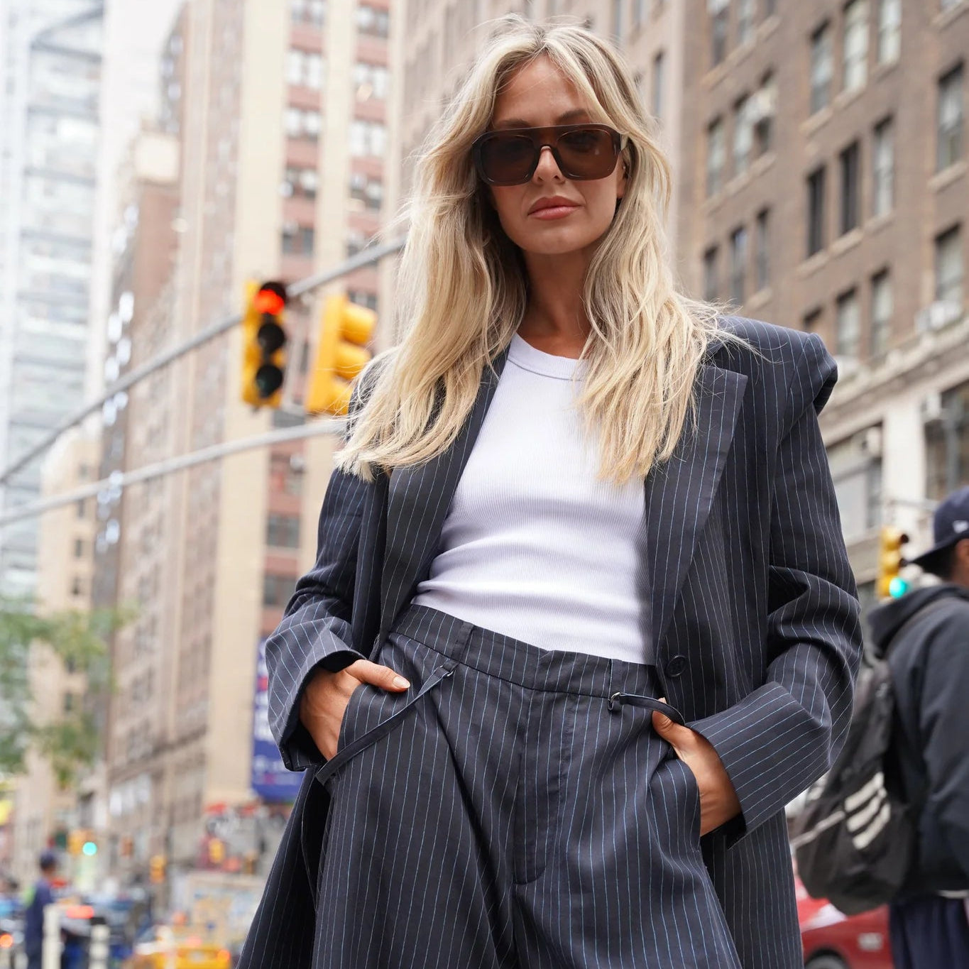 A woman stands on a city street wearing a dark pinstripe blazer and matching wide-leg trousers over a white top, with sunglasses and a blurred urban background.