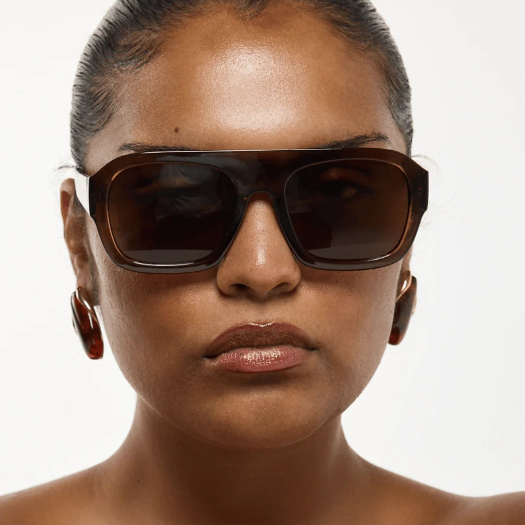 A close-up portrait of a model wearing oversized square sunglasses with dark brown frames and dark lenses, against a light gray background. The brand name 'rhodin.