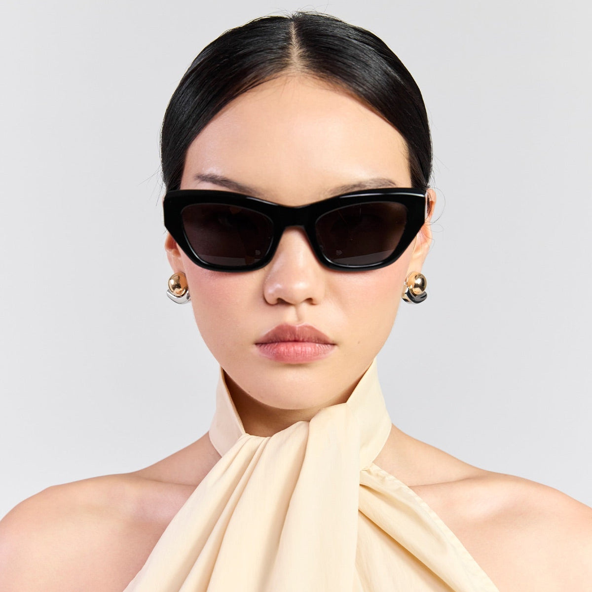 A model with slicked-back black hair wears black cat-eye sunglasses and a draped cream halter top against a light gray studio background.