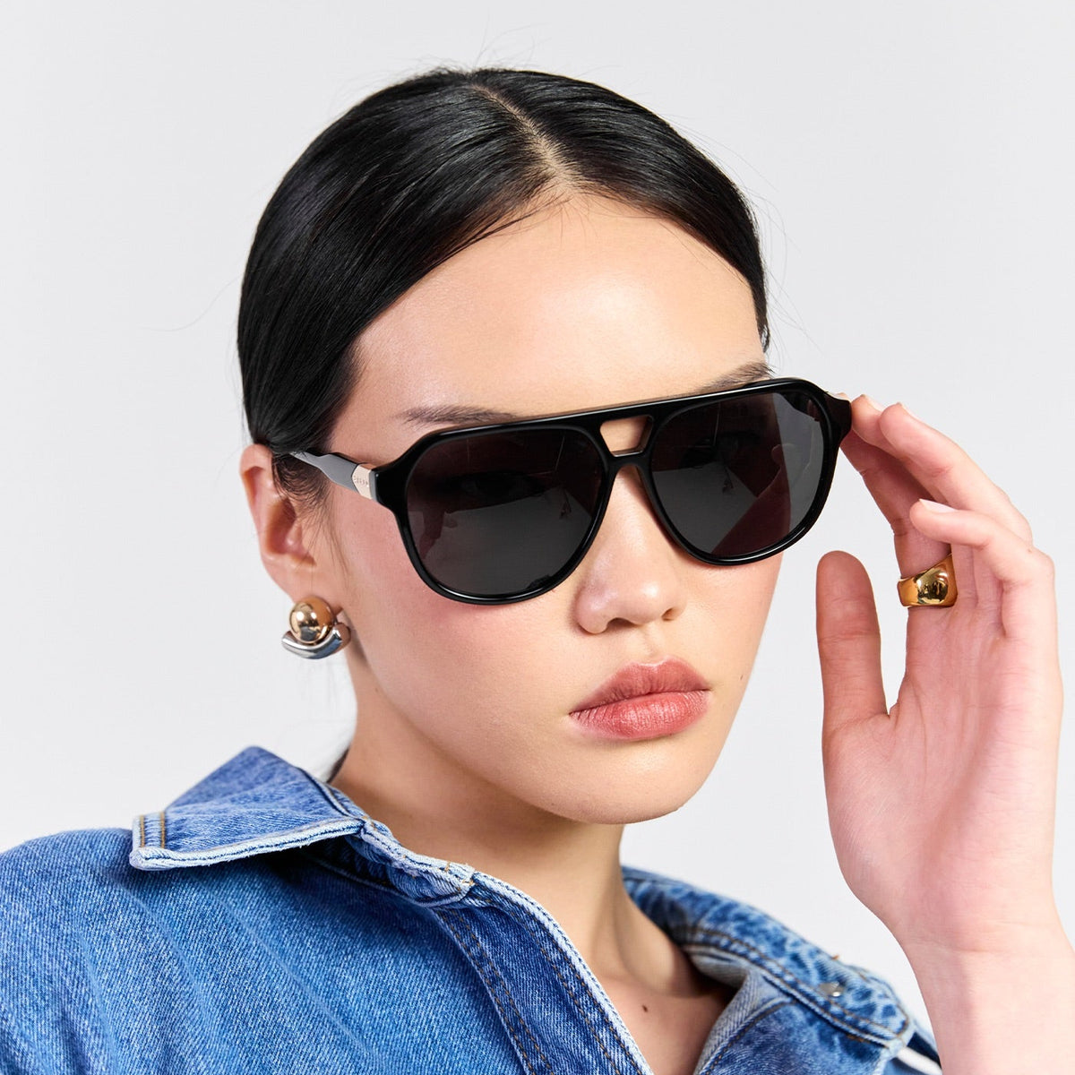 A model wearing black aviator-style sunglasses with a thin silver metal frame and dark lenses, paired with a blue denim shirt against a light gray background.