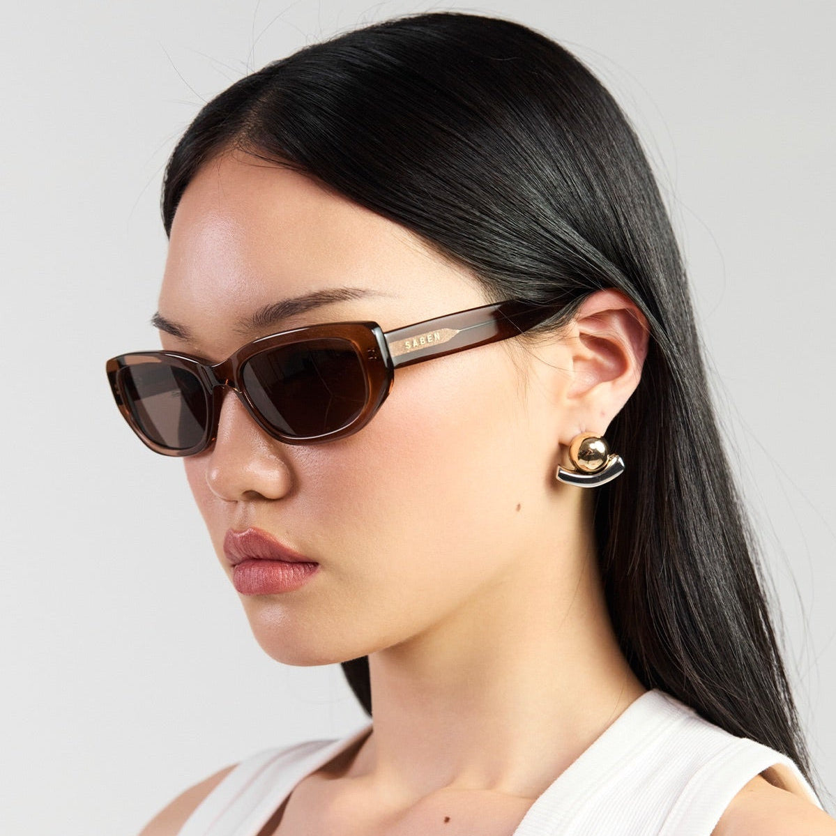 A woman with long black hair is shown in profile wearing dark brown cat-eye sunglasses with gold-tone frames and a white ribbed tank top against a light gray studio background.