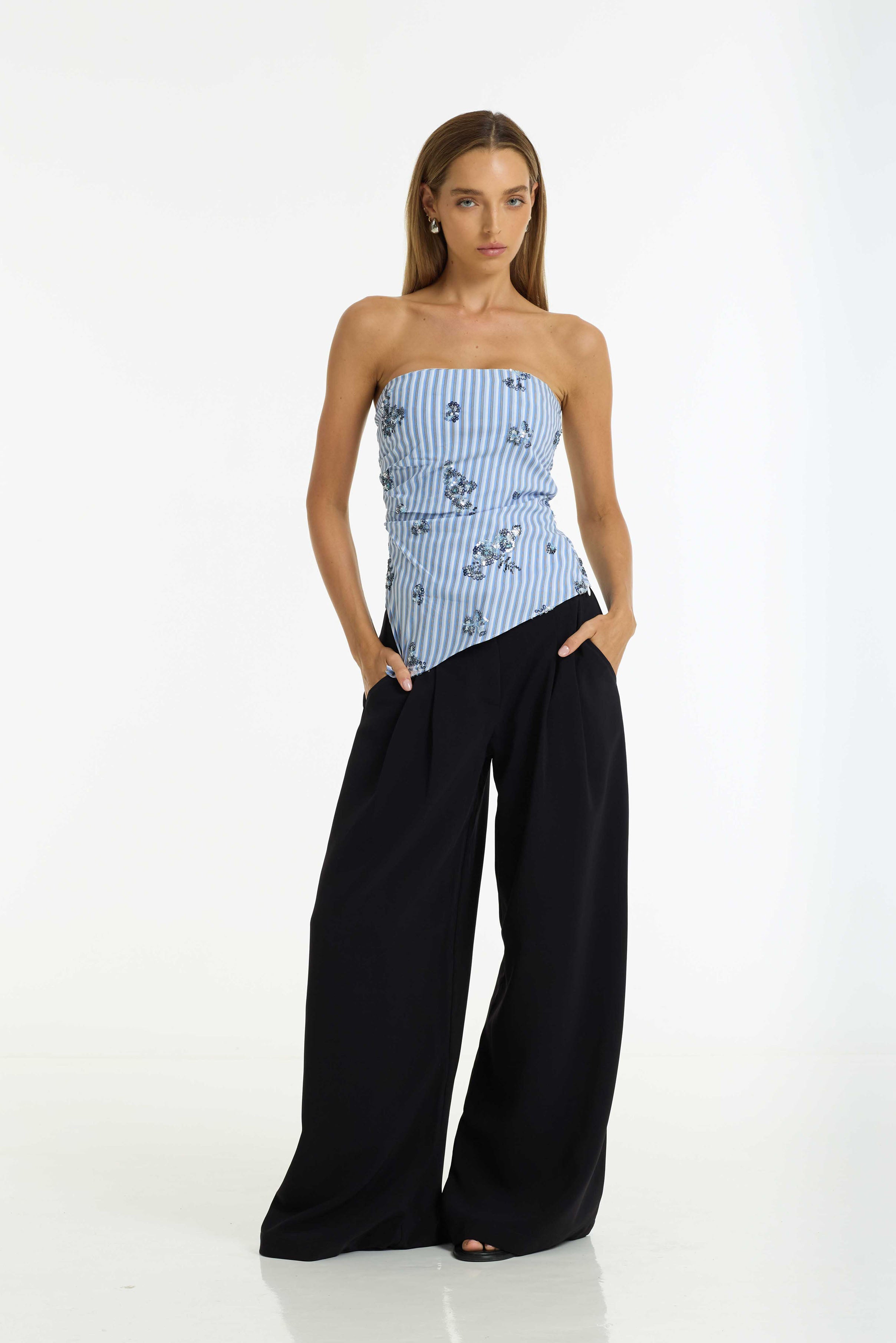 A model wearing a strapless top with vertical blue and white stripes and a delicate floral pattern, paired with wide-leg black pants, against a plain white studio background.