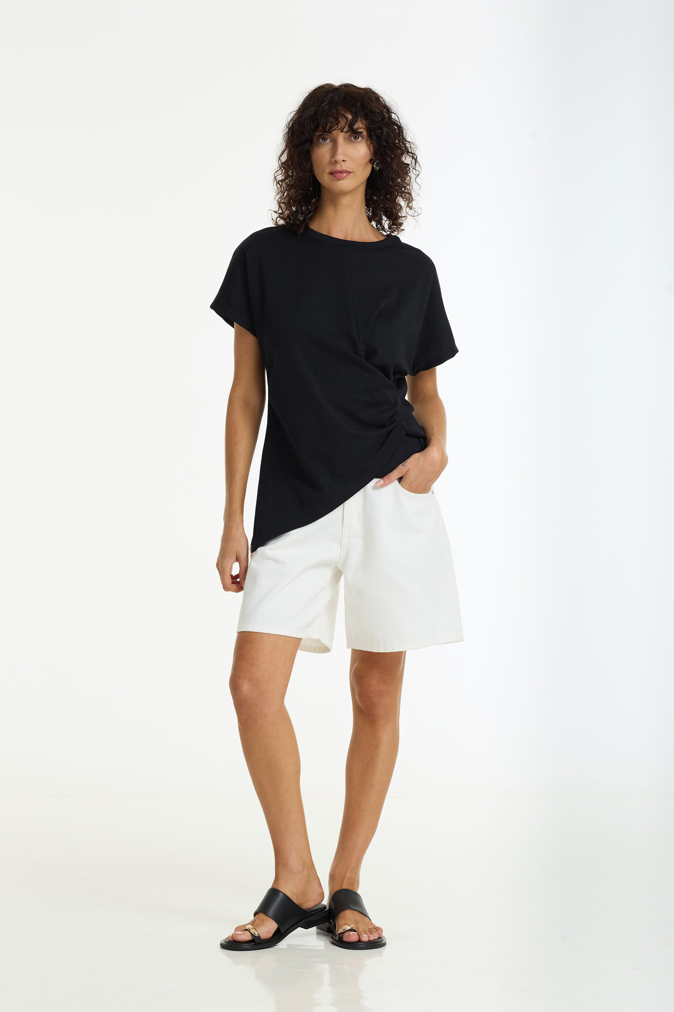 A women wearing a black short-sleeve t-shirt with a twisted side detail, paired with the Sienna Long Denim Shorts in off white by BLAK and black slide sandals, standing against a plain white studio background.