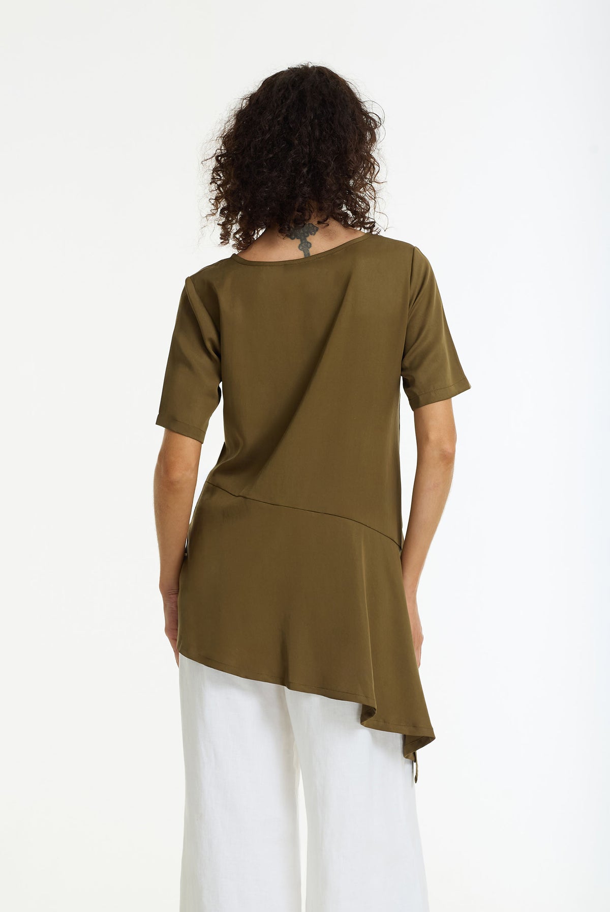 Back view of a woman wearing a khaki soft draped short sleeve top with asymmetric hemline styled with white pants and white pants on a white background