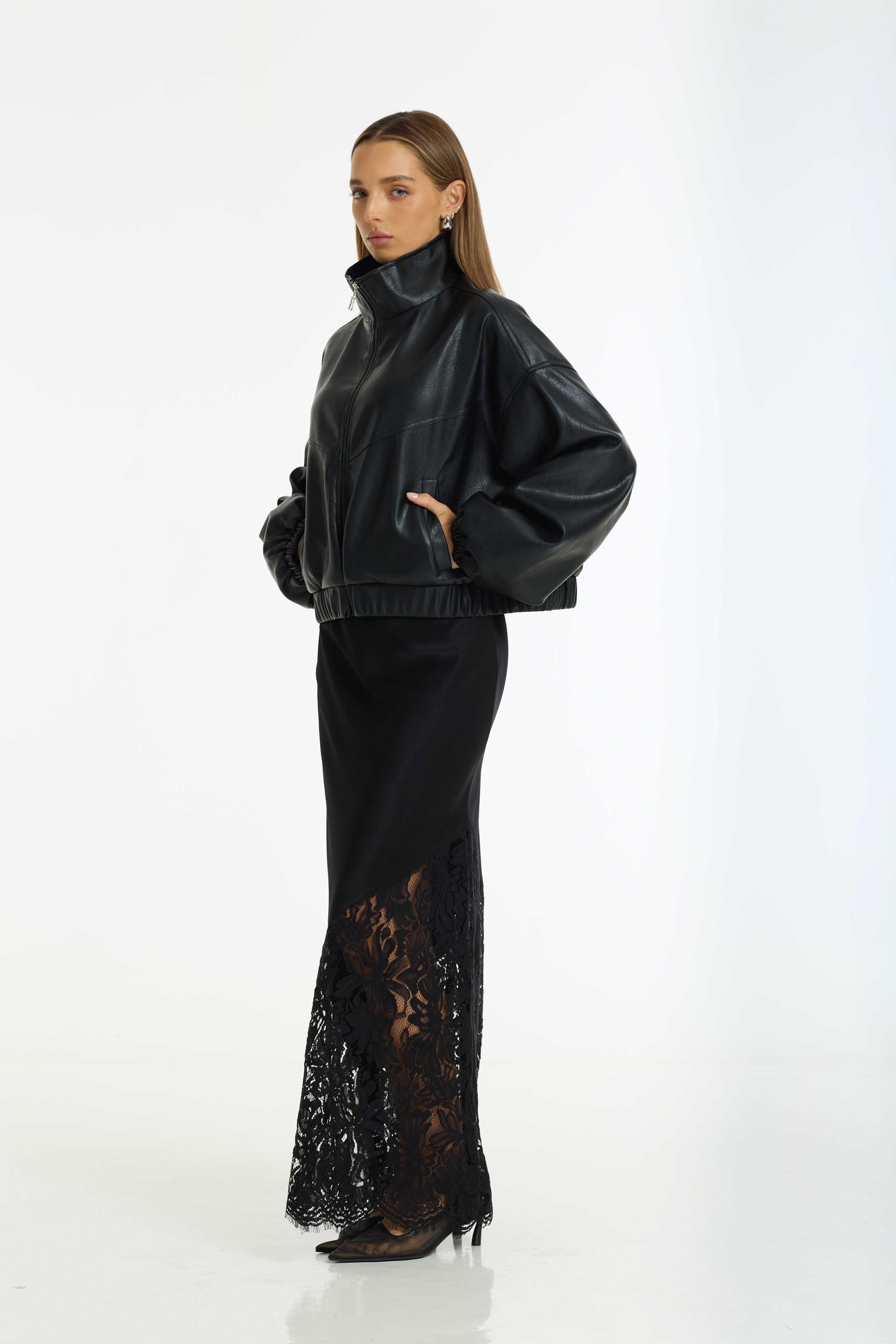 A model stands against a white studio background wearing the Ignite Bomber a black faux-leather cropped zip-up jacket with voluminous sleeves and a Softly Spoken Satin Lace Maxi Skirt in black by BLAK
