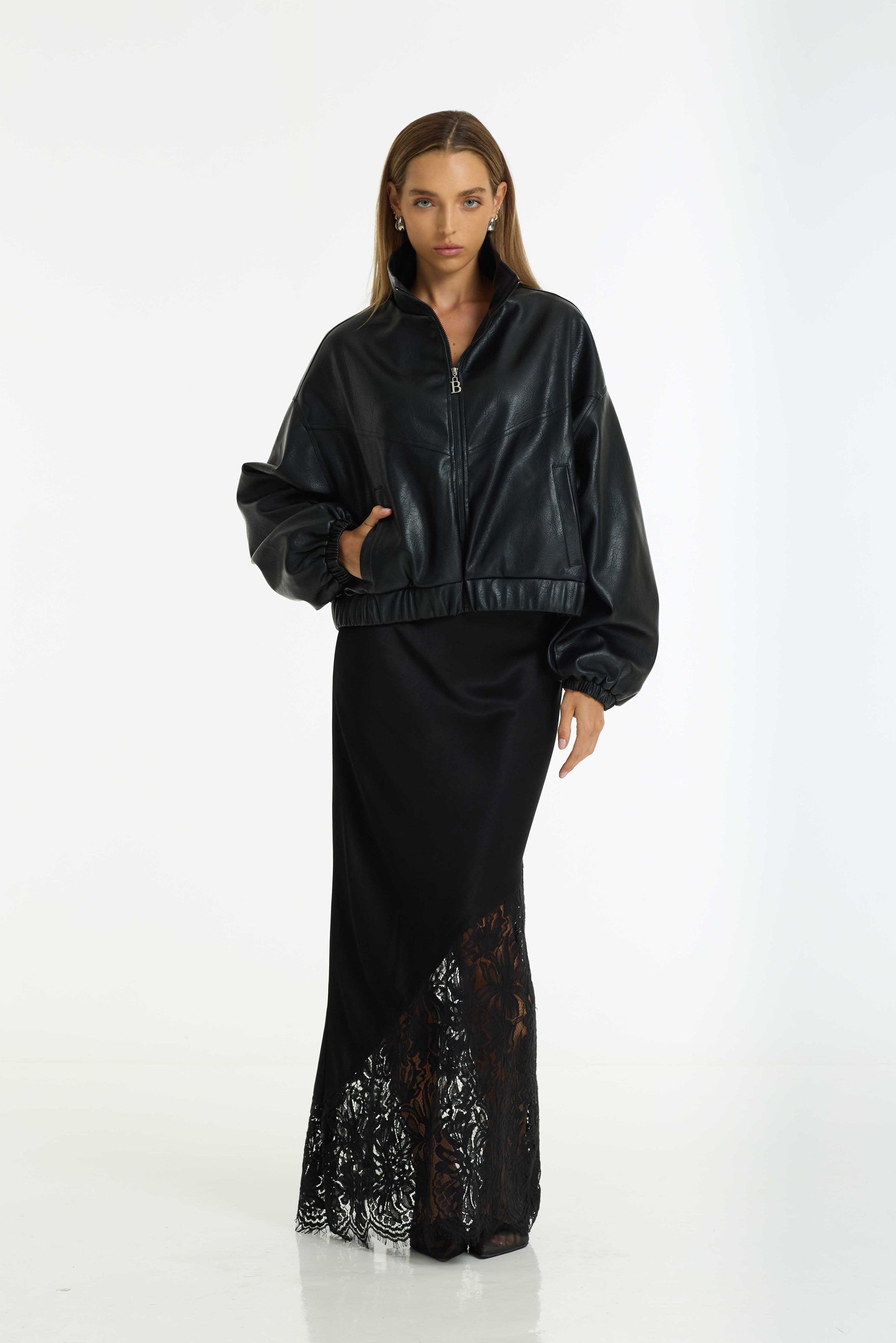 A model stands against a white studio background wearing the Ignite Bomber a black faux-leather cropped zip-up jacket with voluminous sleeves and a Softly Spoken Satin Lace Maxi Skirt in black by BLAK