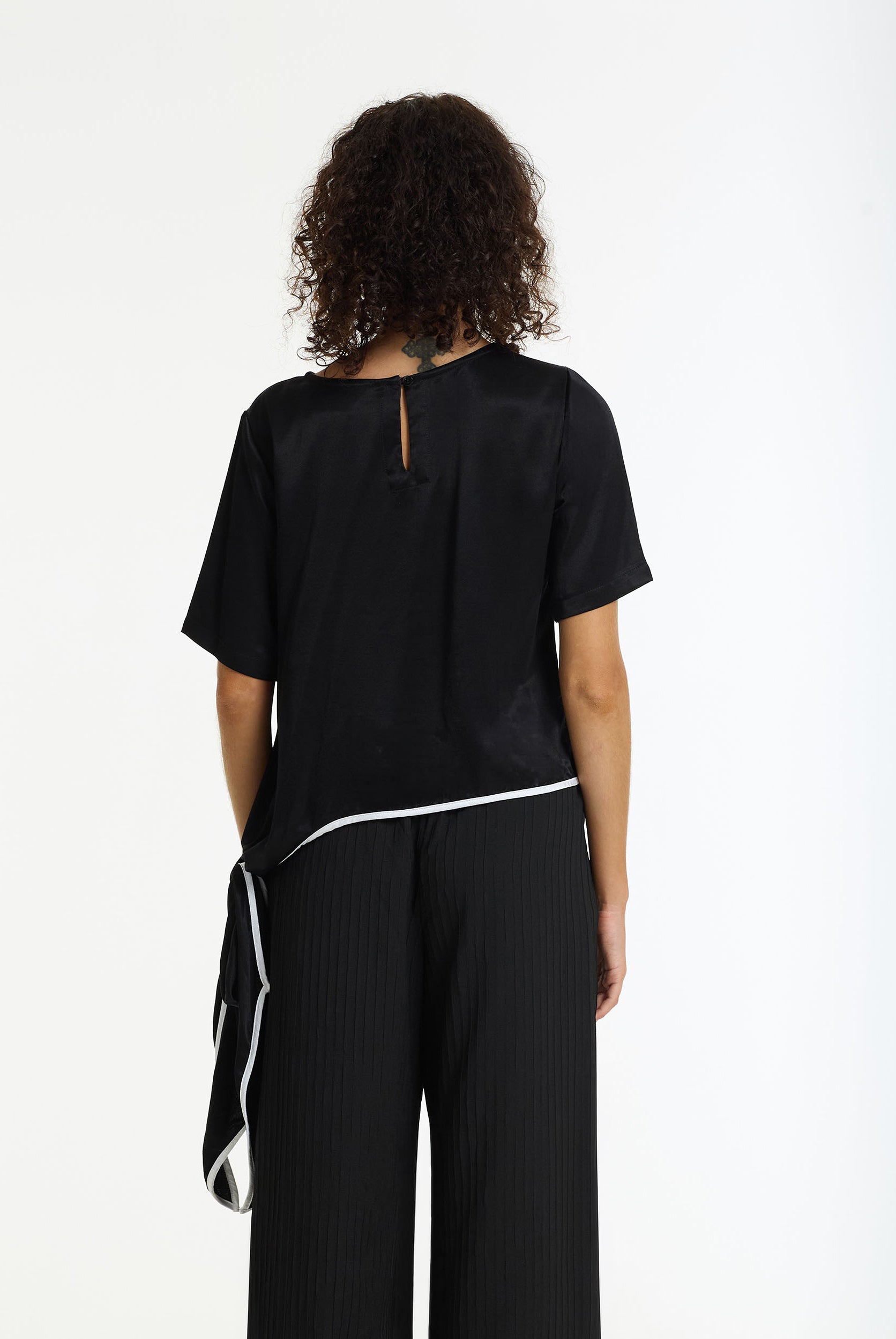 Back view of a women wearing The Knot Satin Tie Top black white binding by BLAK and black trouser with a white background