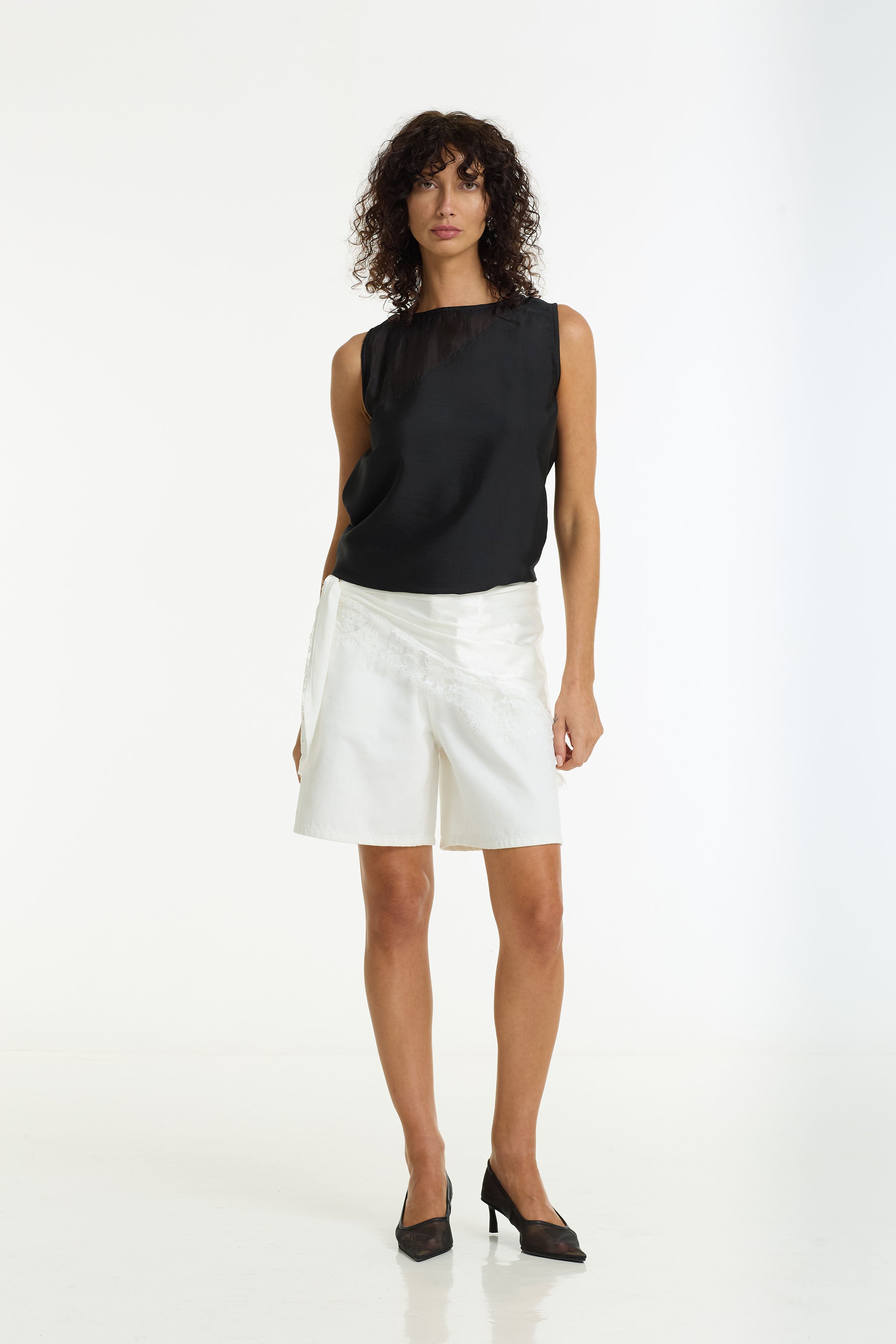 A model stands against a white studio background wearing a sleeveless black top with a sheer yoke and white shorts, paired with The Lace Edit Bandana Scarf in ivory by BLAK.