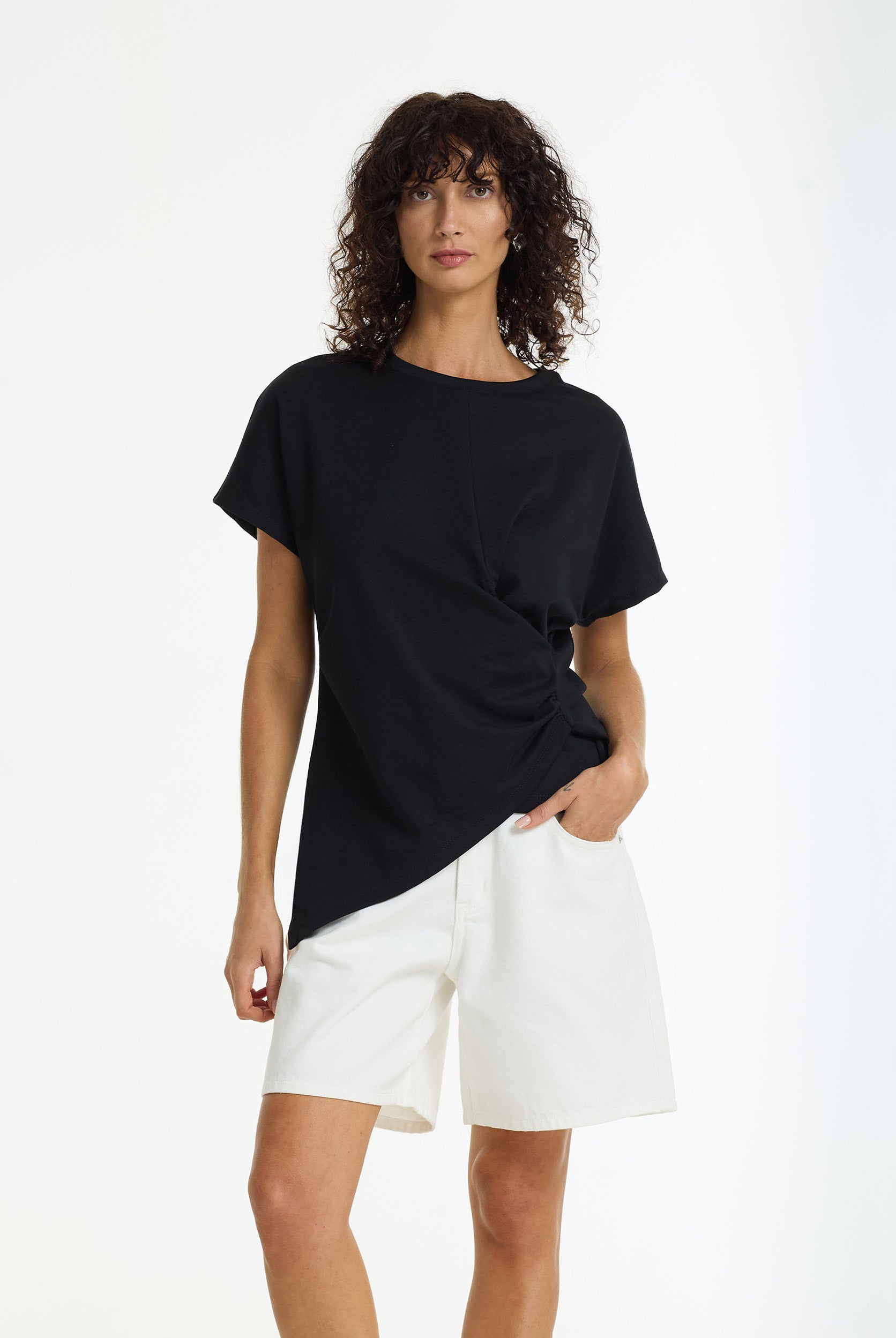 Woman wearing the Time Slows Asymmetric Tee black cotton t-shirt by BLAK and white shorts on a white background