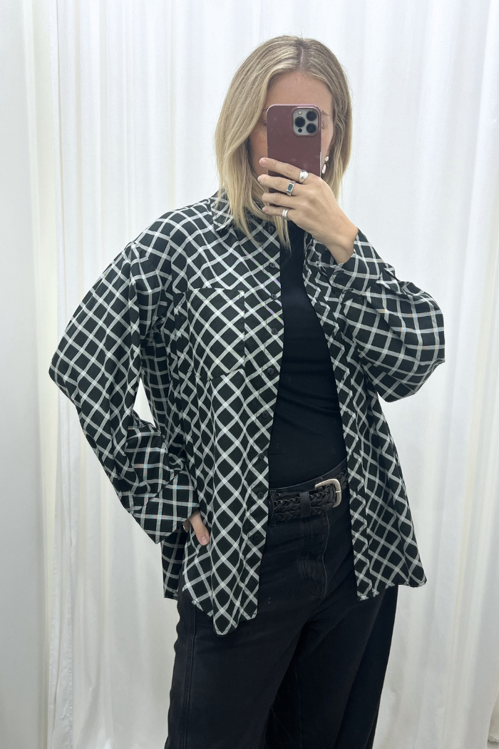 A woman taking a mirror selfie wearing the Two Makes One Oversized Cotton Shirt black white cross hatch by BLAK over a black top and black pants, set against a white curtain background.