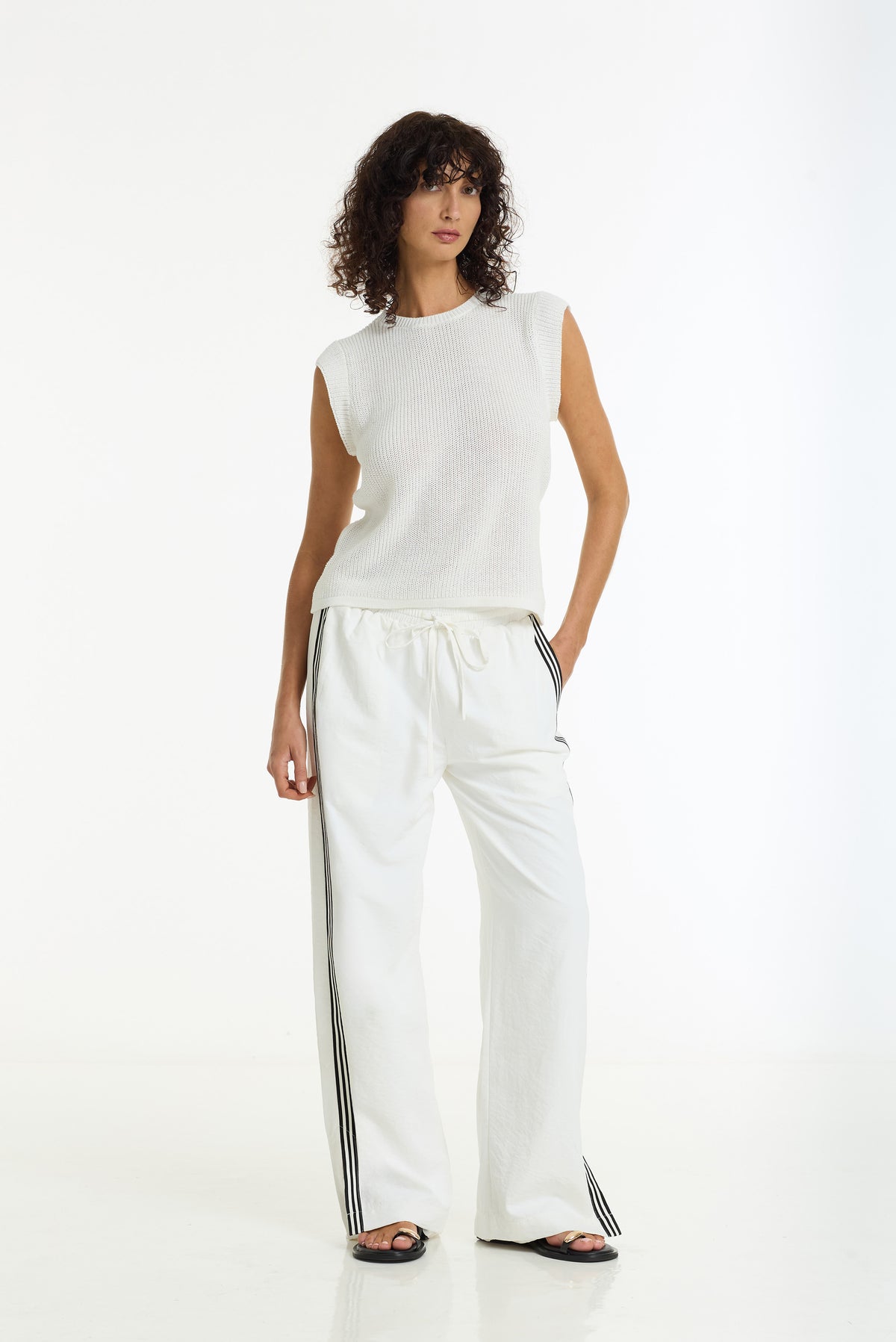 A model wearing a white sleeveless knit top and matching white wide-leg pants with black side stripes, standing against a plain white studio background.
