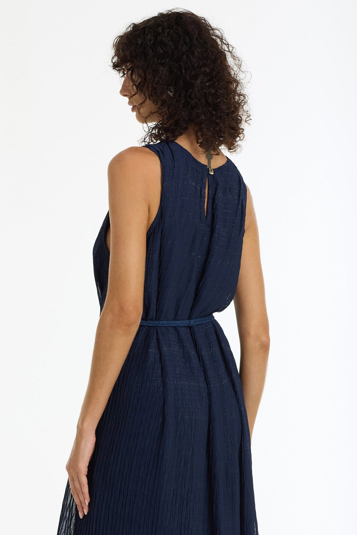 A woman modeling a sleeveless navy blue maxi dress from the back, featuring a tie-back neckline, a matching waist tie, and a flowing skirt, set against a plain white studio background.