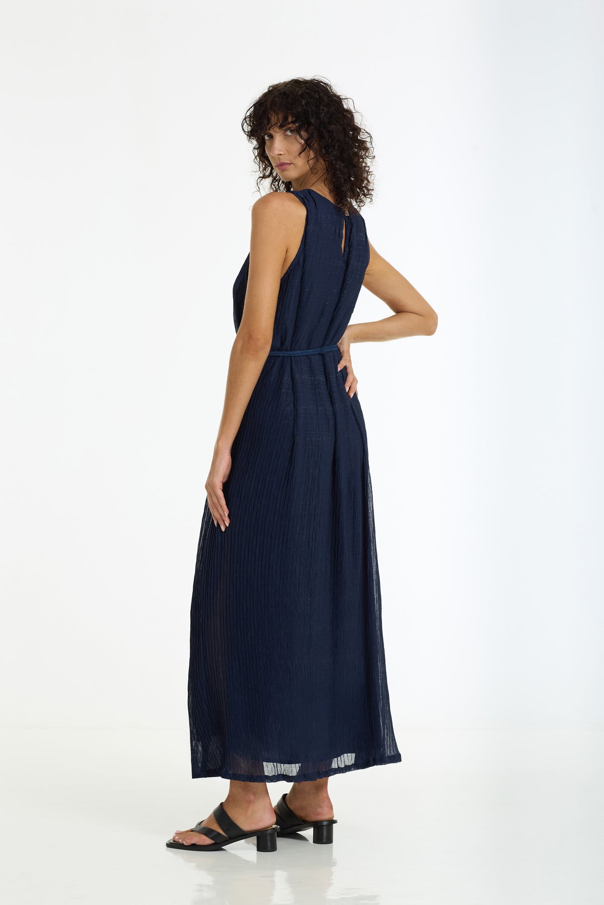 A woman modeling a sleeveless navy blue maxi dress with a pleated texture, shown from a side-back angle against a clean white studio background.