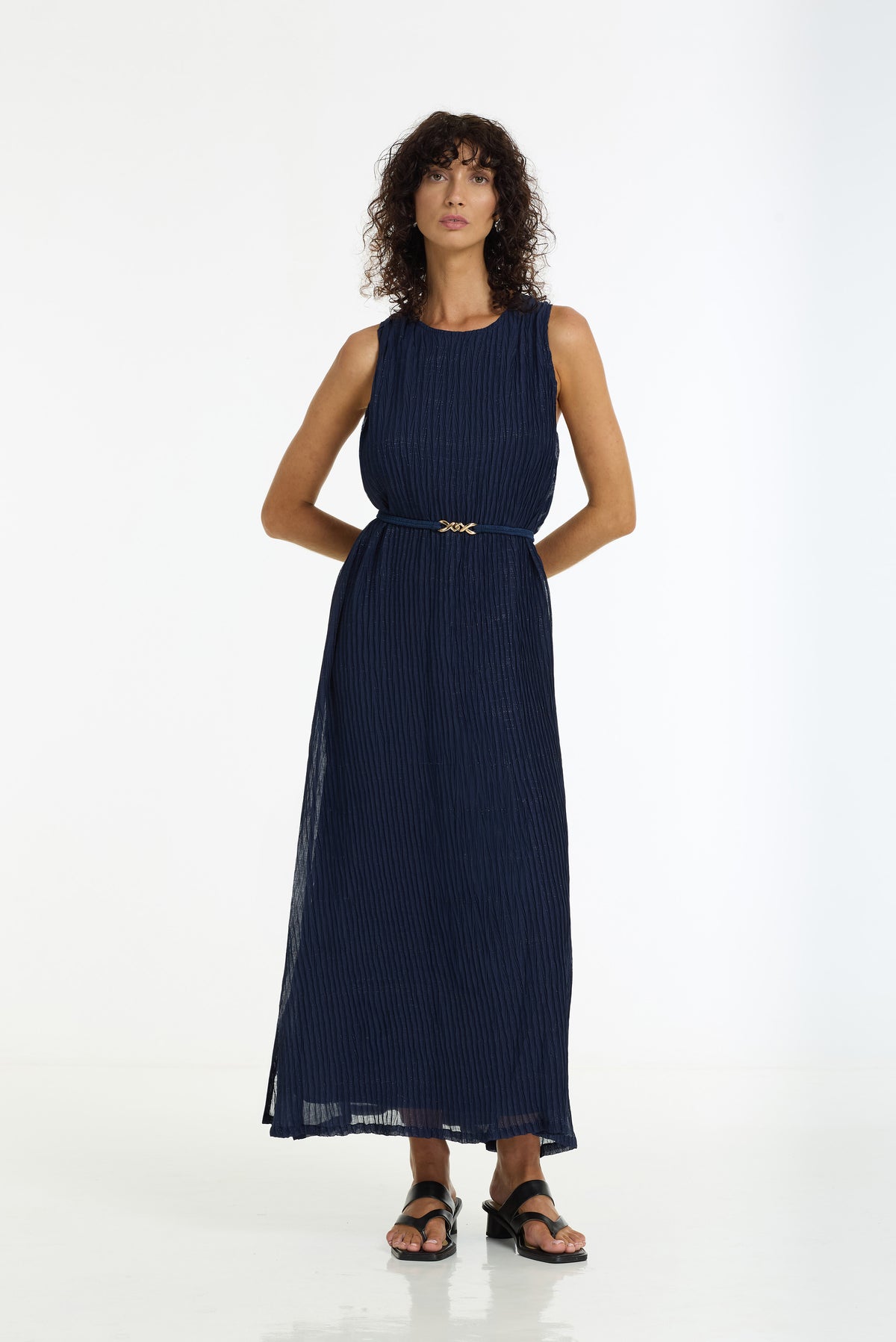 A woman modeling a sleeveless navy blue pleated maxi dress with a matching belt featuring a small gold-tone bow, set against a plain white studio background.