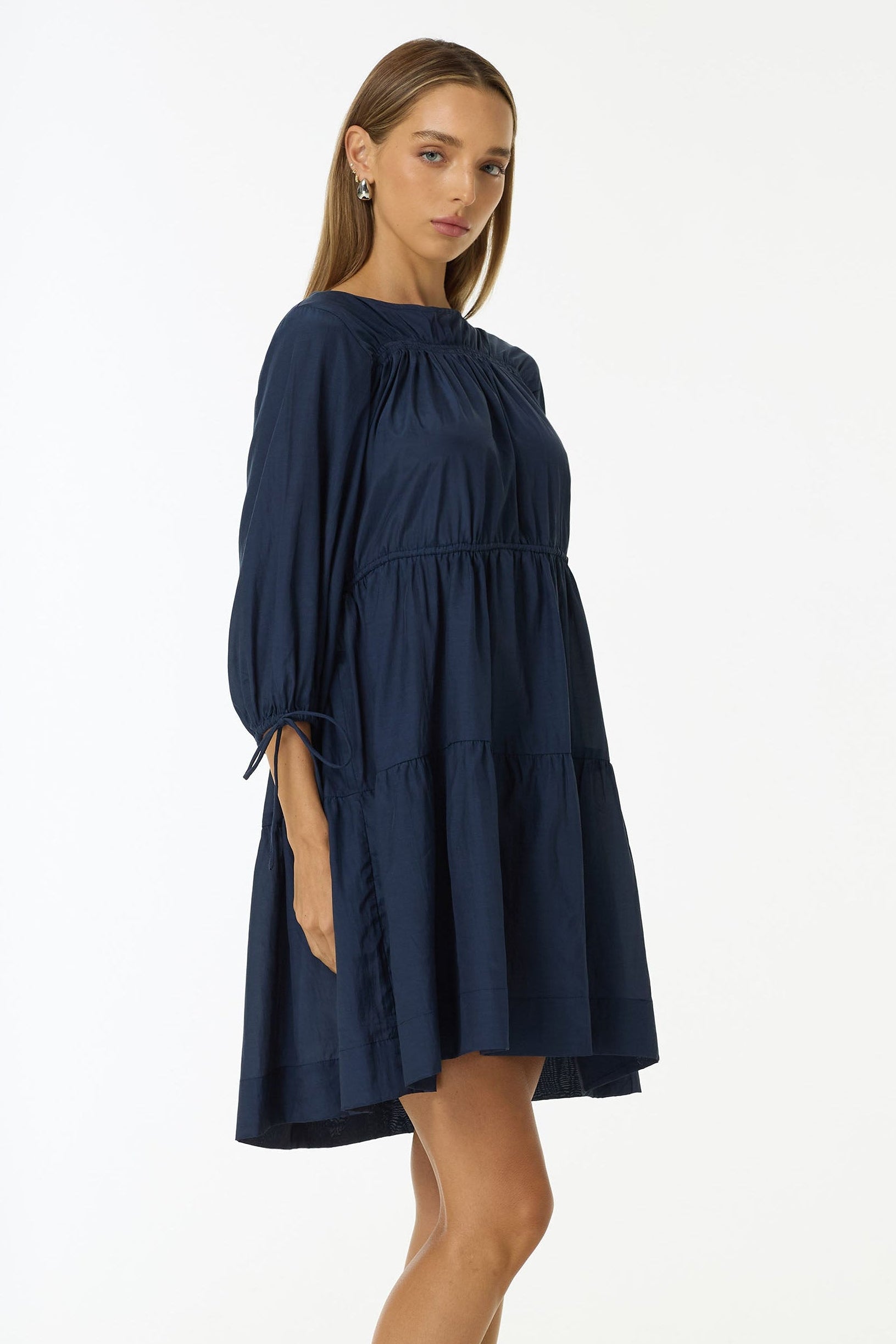 A woman modeling a navy blue tiered mini dress with long puff sleeves and black flip-flop sandals, standing against a plain white studio background.