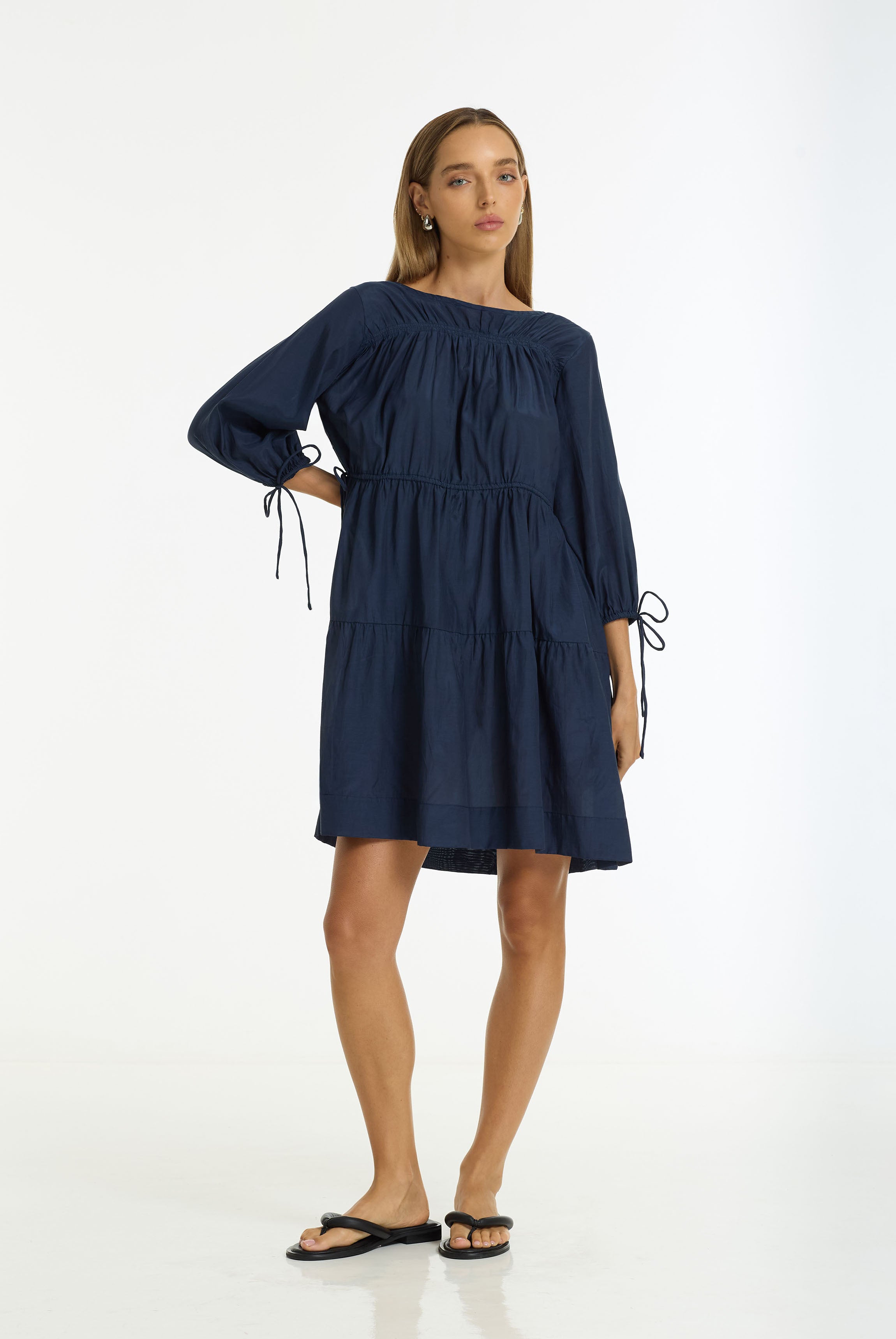 A woman modeling a Where Hearts Fall Cotton Voile Dress in navy by BLAK, paired with black flip-flop sandals against a plain white studio background.