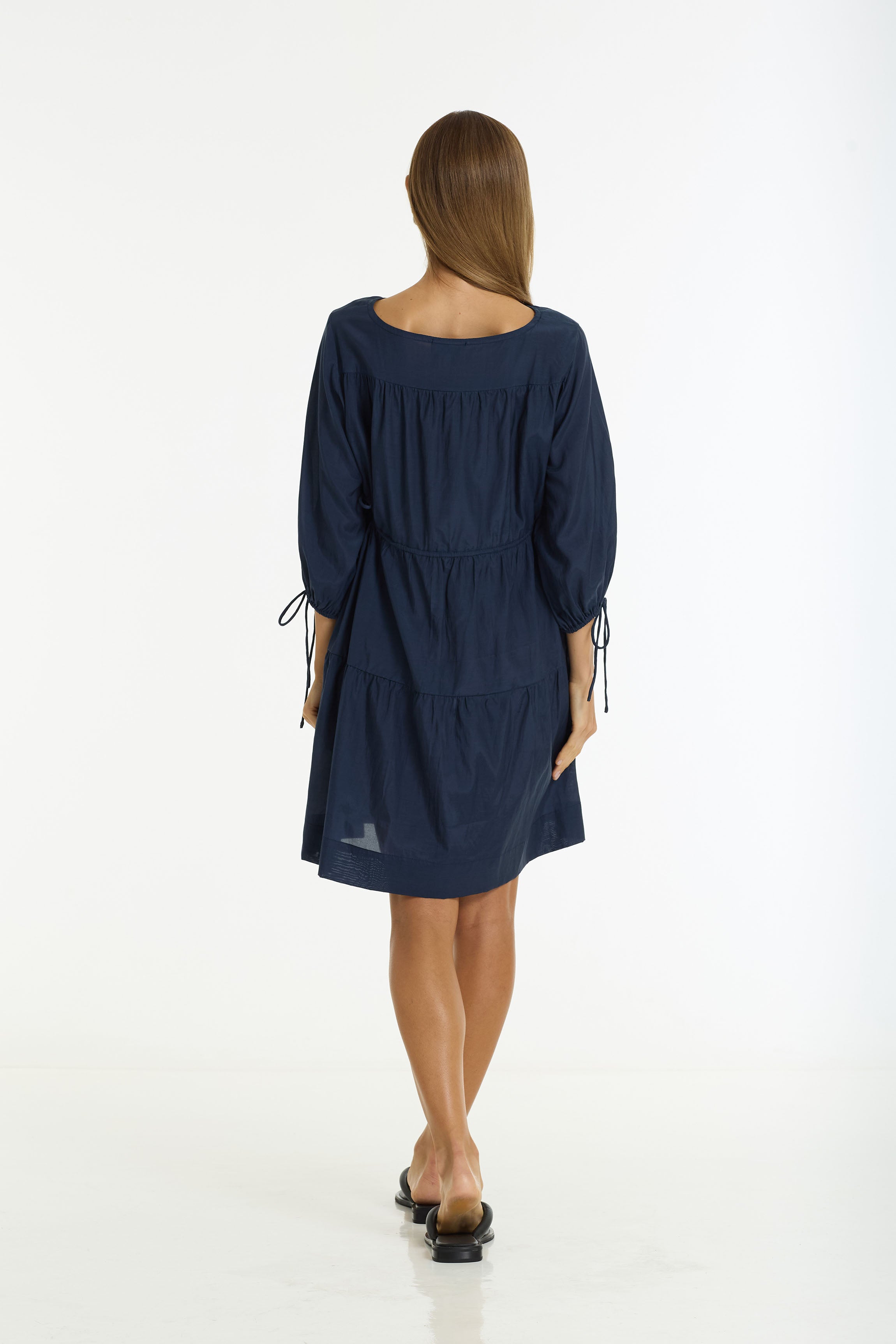 A woman viewed from the back wearing a navy blue tiered dress with three-quarter sleeves, side tie details, and black flat sandals, set against a plain white studio background.